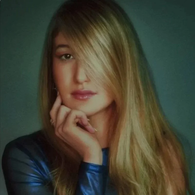 A woman with long blonde hair partially covering one eye, wearing a dark top, and resting her chin on her hand, against a dark background.