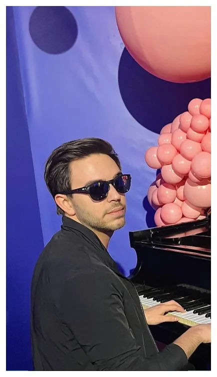 A man wearing sunglasses and a black jacket playing a piano, with pink and purple balloons on a blue wall background.