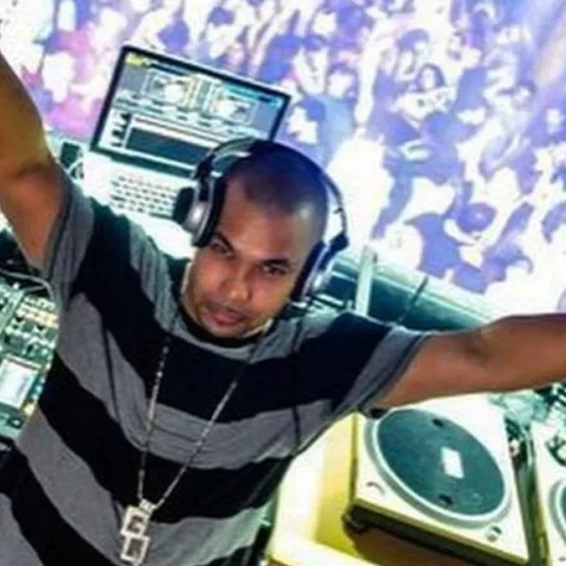 DJ wearing headphones with arms outstretched, surrounded by DJ equipment including turntables and a laptop, at a party or club.