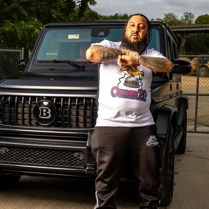A man with tattoos and a beard standing in front of a black luxury SUV, wearing a white T-shirt with cartoon graphics and black pants.