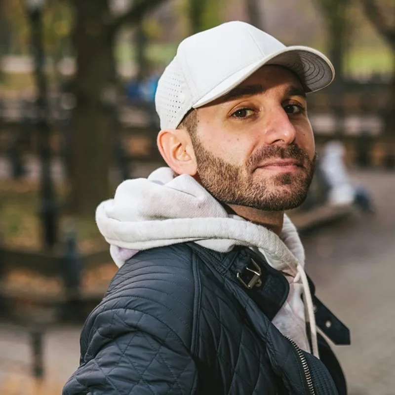 A man wearing a white baseball cap, gray hoodie, and black quilted jacket outdoors in a park with blurred trees and a bench in the background.
