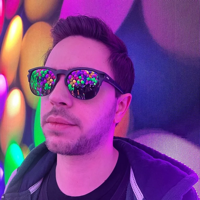 A man wearing black sunglasses reflects colorful bright lights. He has short dark hair, light facial hair, and is dressed casually with a black shirt and hoodie. The background features vivid, colorful, abstract lighting.