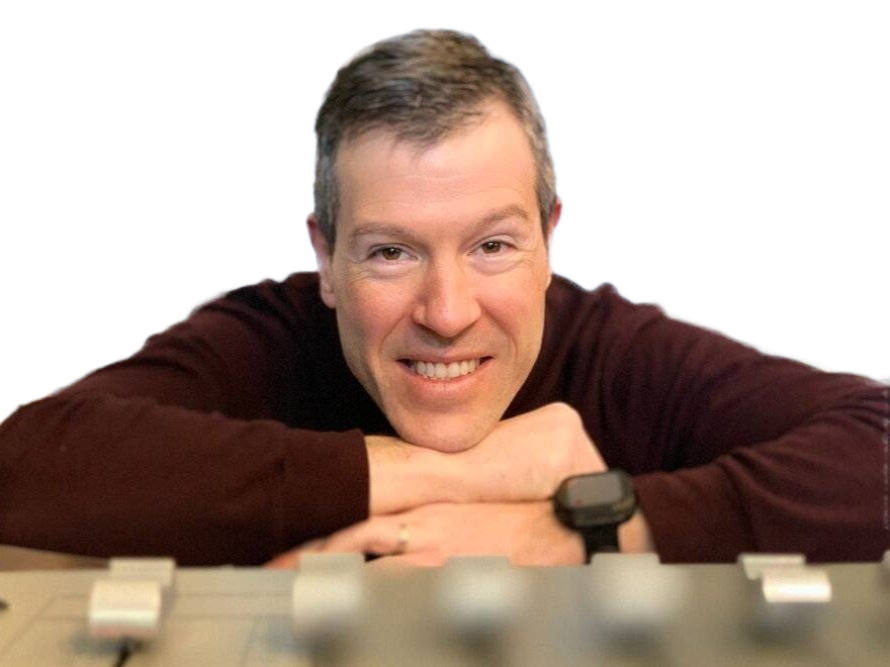A smiling man with short brown hair leaning on a table, wearing a dark maroon shirt and a wristwatch.