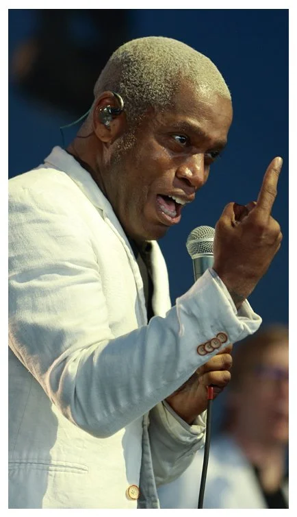 A man with dyed blond hair speaking into a microphone, pointing his finger upward, wearing a white blazer, on stage.