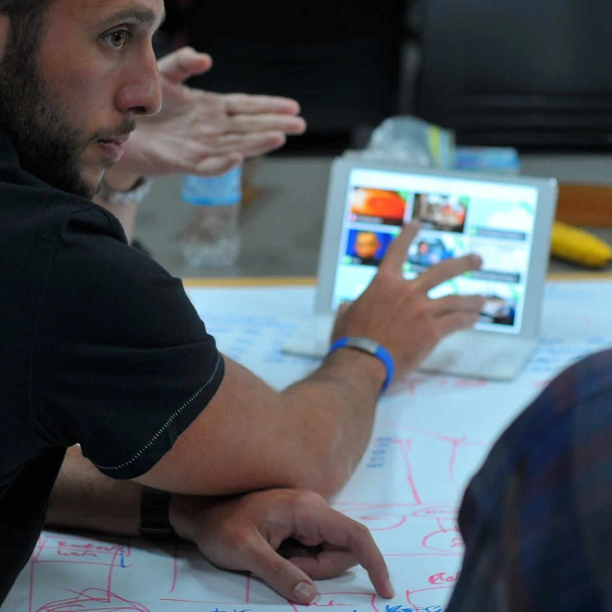 Man pointing to a tablet screen with a presentation or video thumbnails at a meeting.