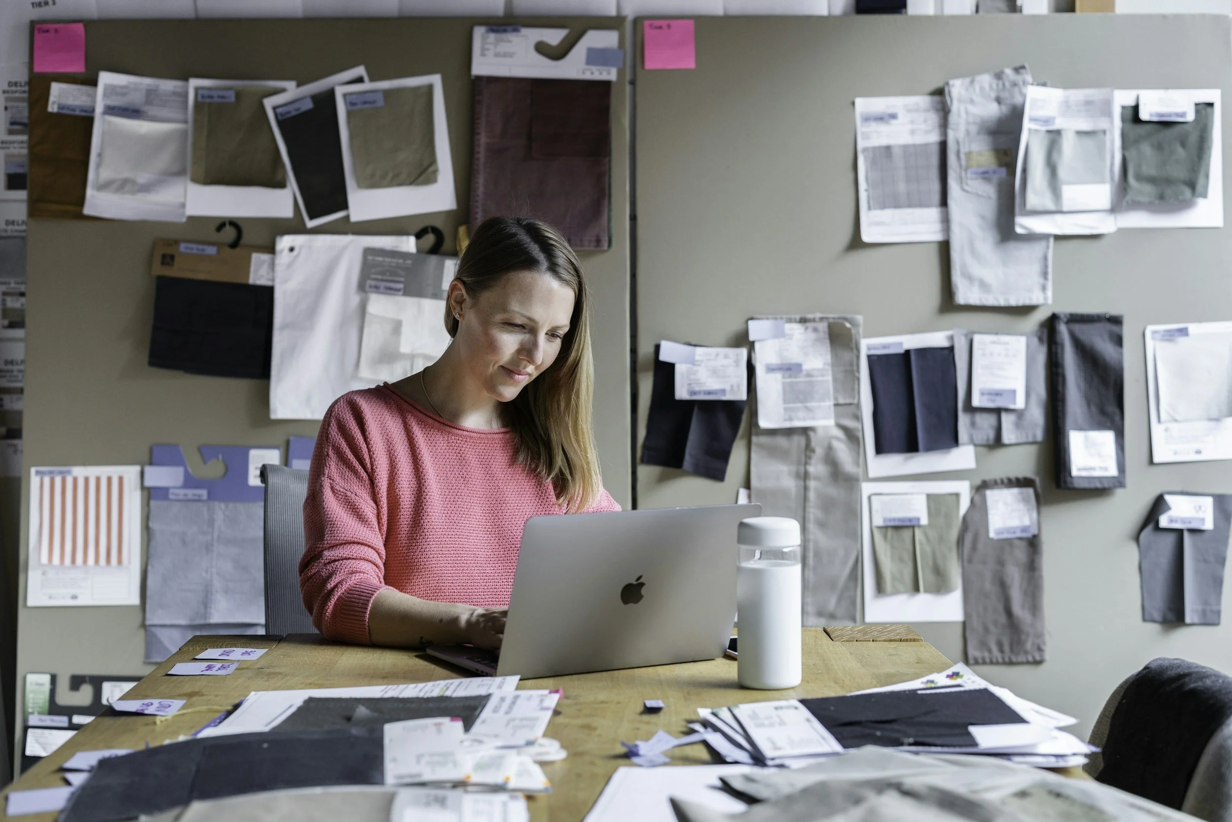 A woman with long brown hair sitting at a desk working on a laptop in a creative workspace. The background features a wall with fabric swatches, color samples, and design plans pinned up.