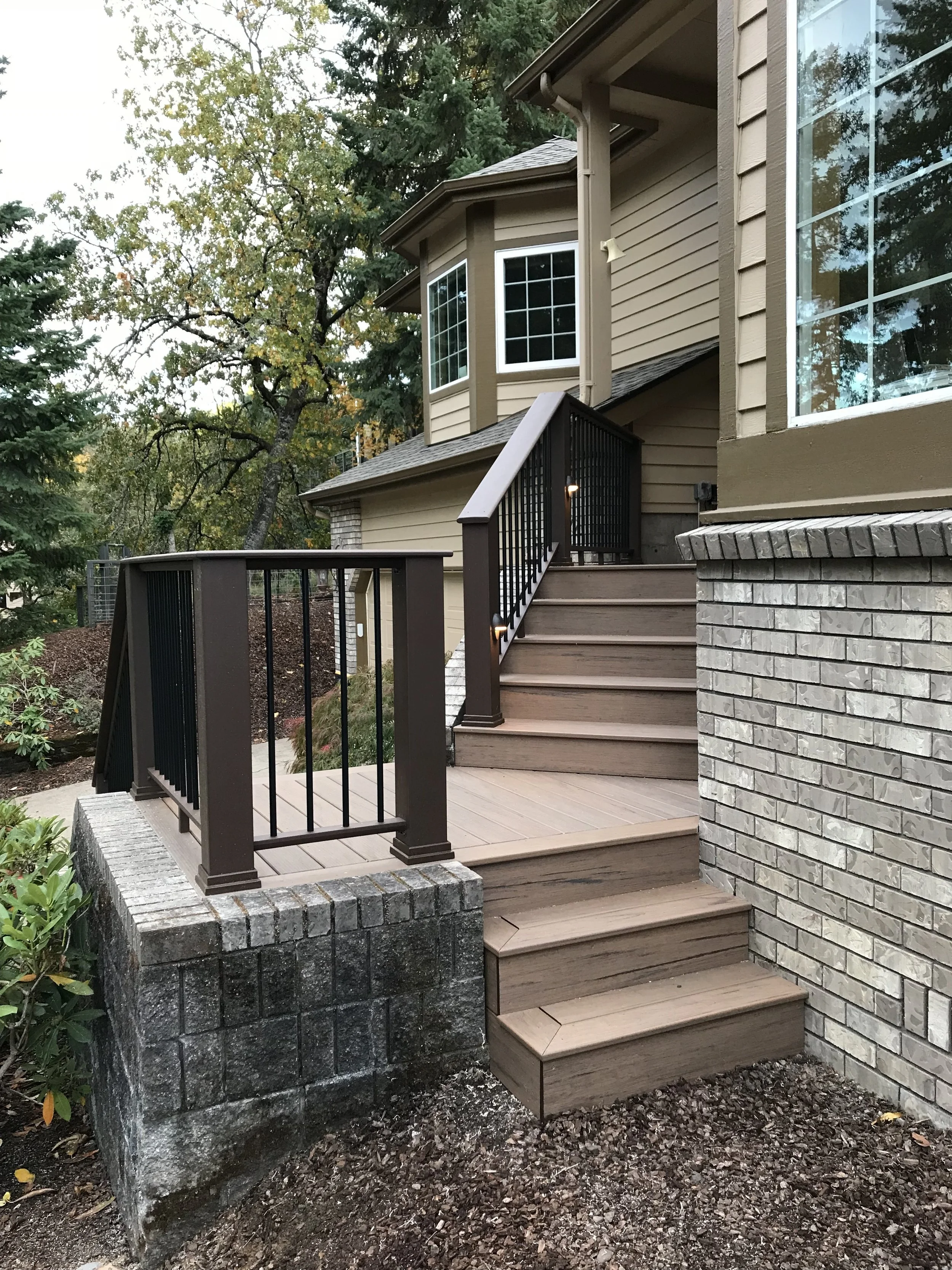 A backyard wooden deck with stairs, black metal railing, and a brick foundation next to a house with beige siding and large windows surrounded by trees.
