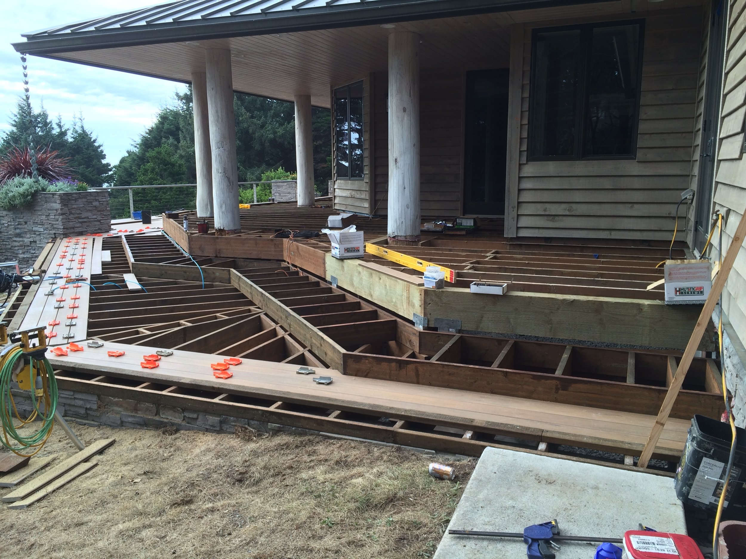 Construction site of a house deck with wooden framing, construction tools, and materials around, with a house with wood siding and trees in the background.