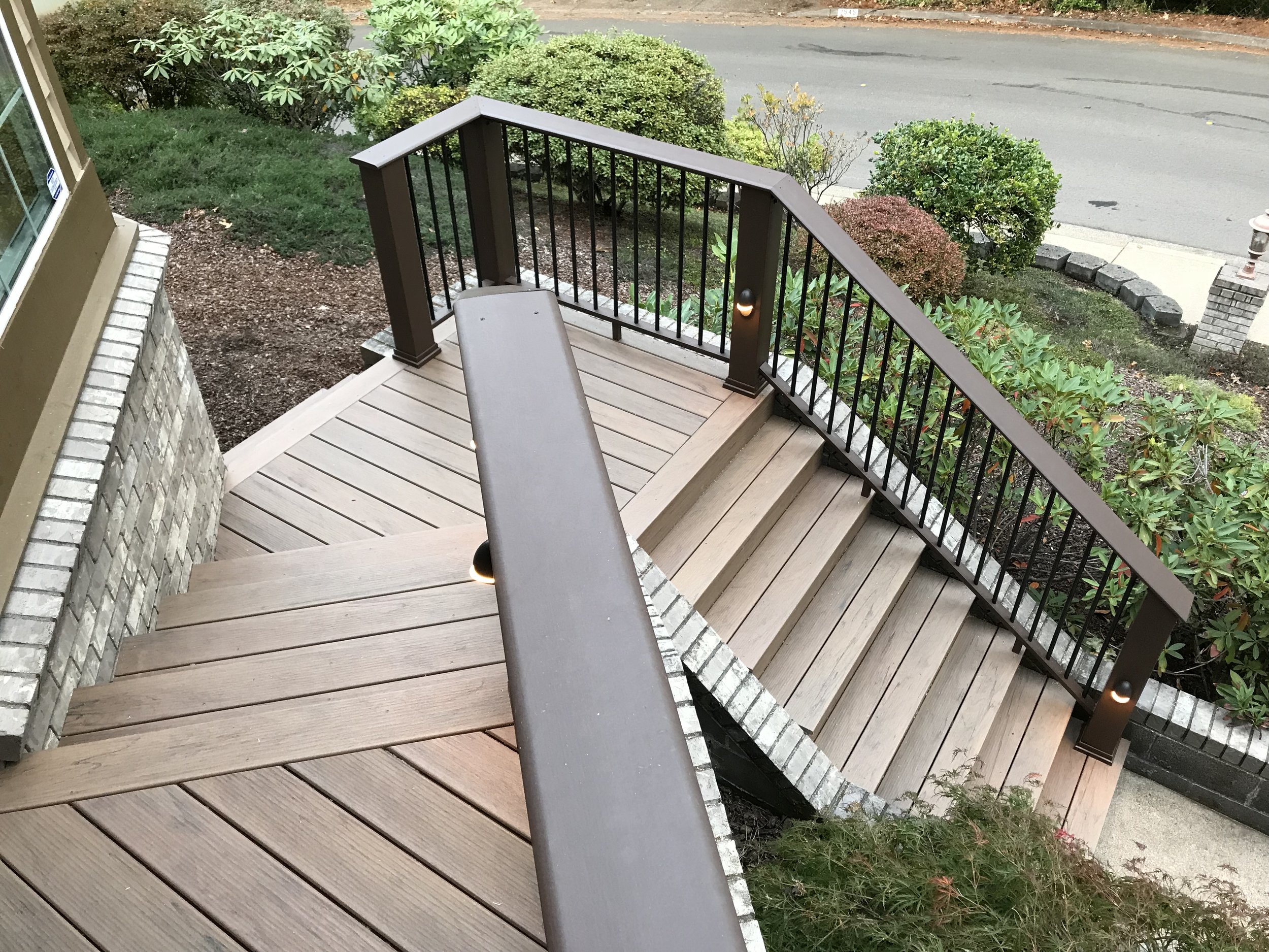 View of a small wooden front porch with a brown railing and a single bench, surrounded by bushes and plants, with a concrete sidewalk and street in the background.