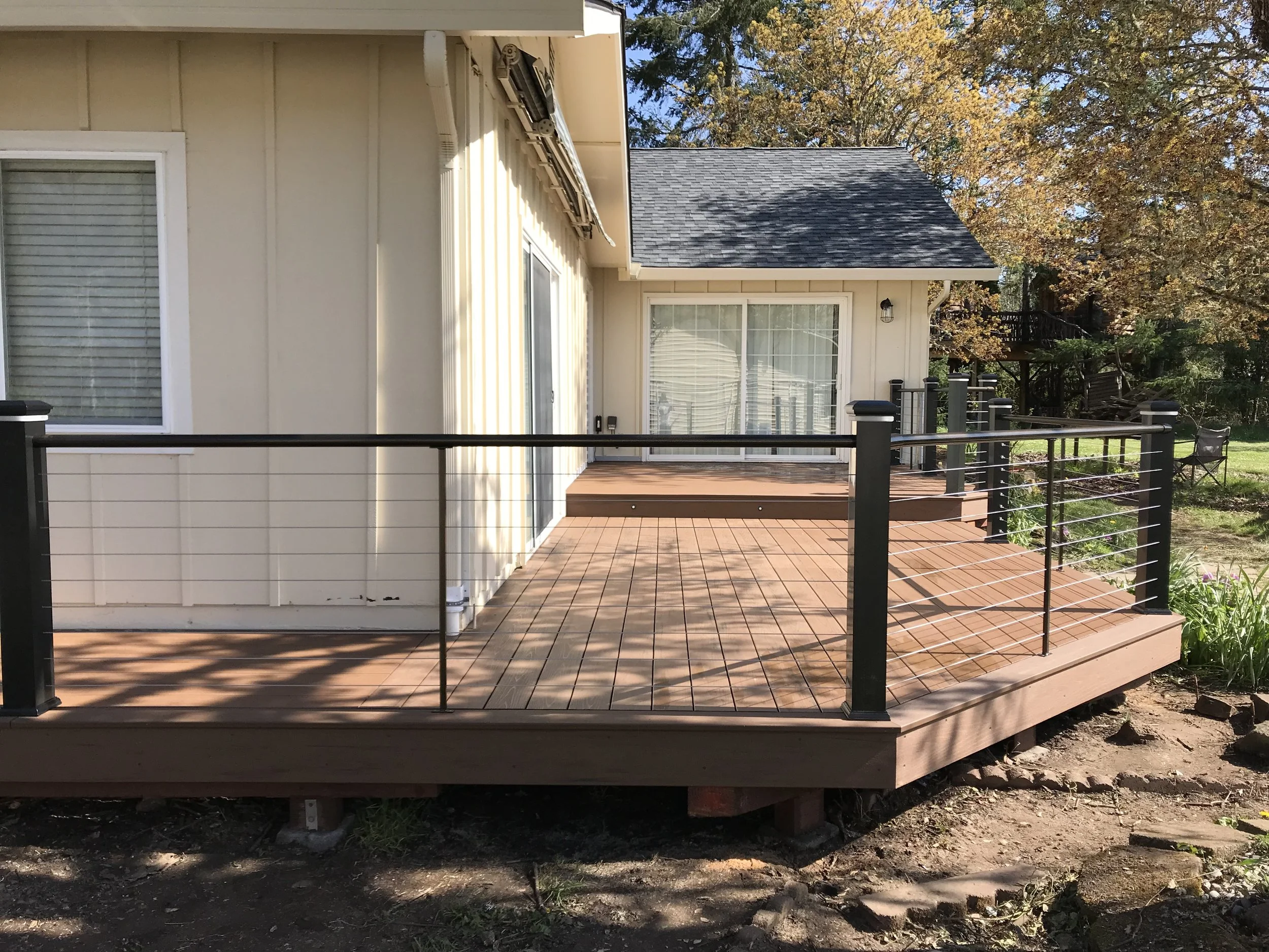 A backyard deck with black metal railing, beige wooden flooring, and outside walls of a house with beige siding, two windows, and a sliding door. An outdoor light fixture is mounted on the house. Trees with autumn leaves are visible in the background