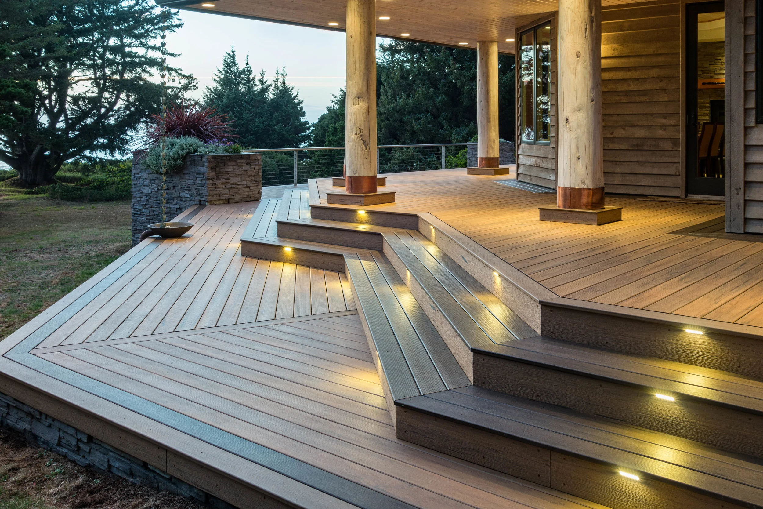 Wooden deck with stairs and built-in lights, attached to a house with wood siding, surrounded by trees and greenery, during twilight.