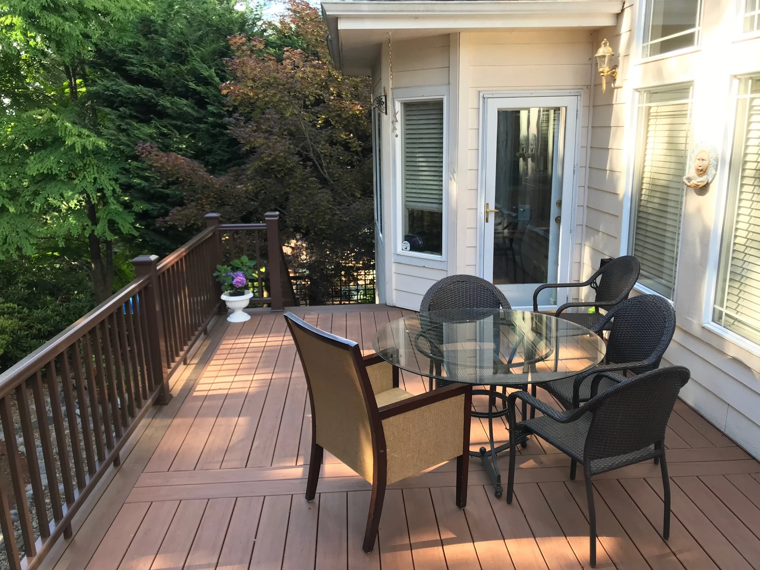 A backyard patio with a wooden deck, outdoor table with glass top, surrounded by chairs, white house siding, a sliding glass door, a window with blinds, a house porch light, a decorative mask, a potted plant, and lush green trees in the background.