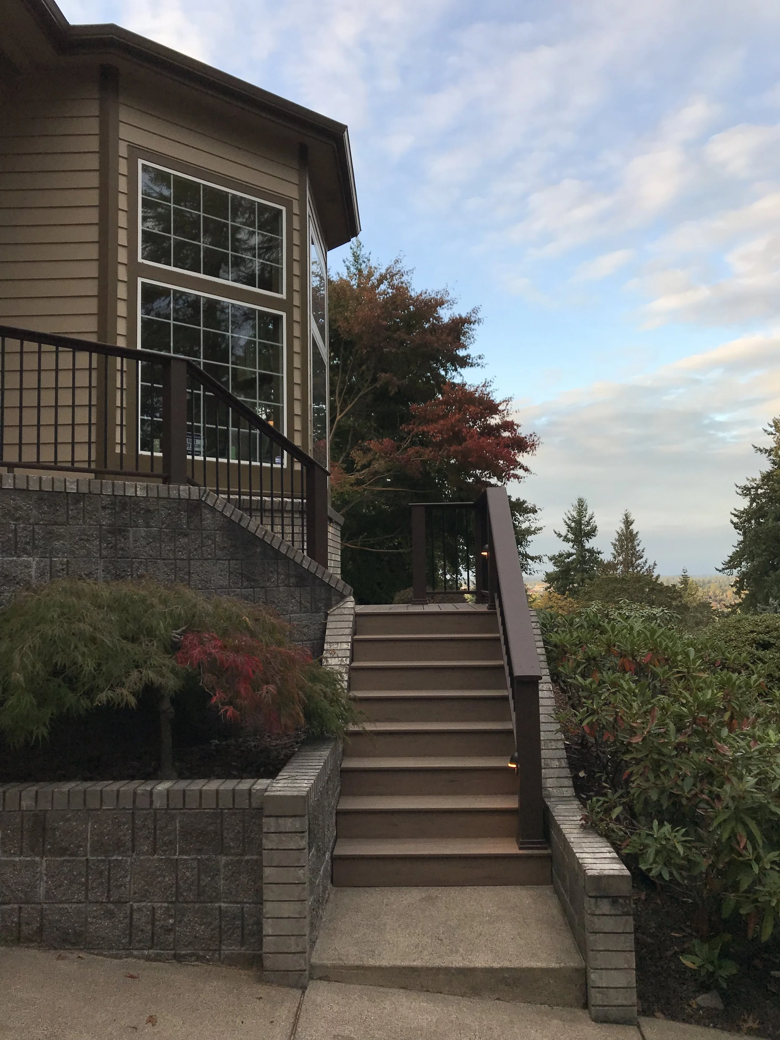 A staircase with dark brown rails and beige steps leads up to a house with beige siding and large windows. There is landscaping with shrubs and trees, some with red leaves, and a cloudy sky in the background.