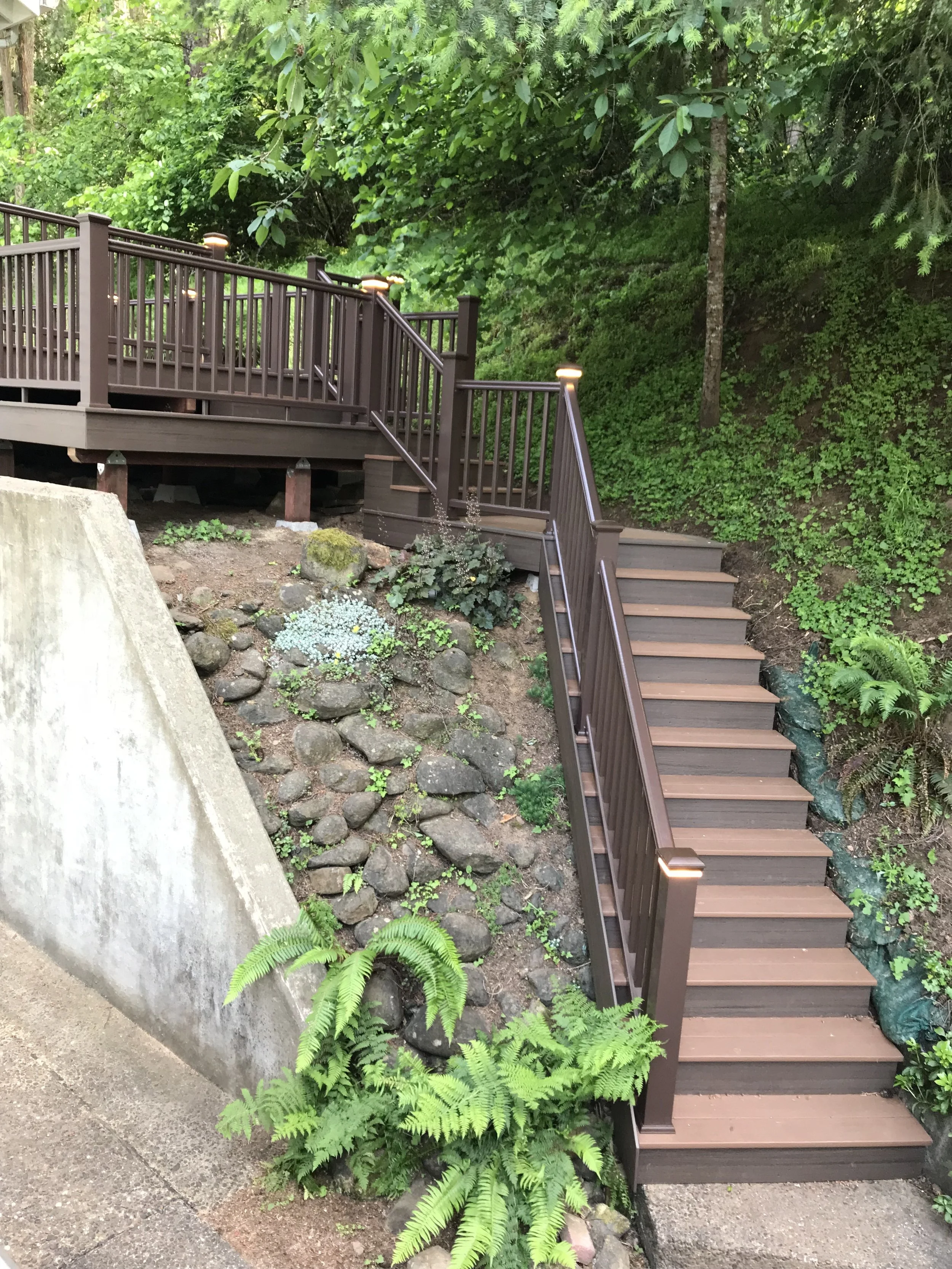 A wooden staircase with a railing leading down to a garden area with lush green plants and trees. A small patch of decorative rocks with plants is visible at the base of the stairs. There is a concrete barrier or wall to the left side of the image.