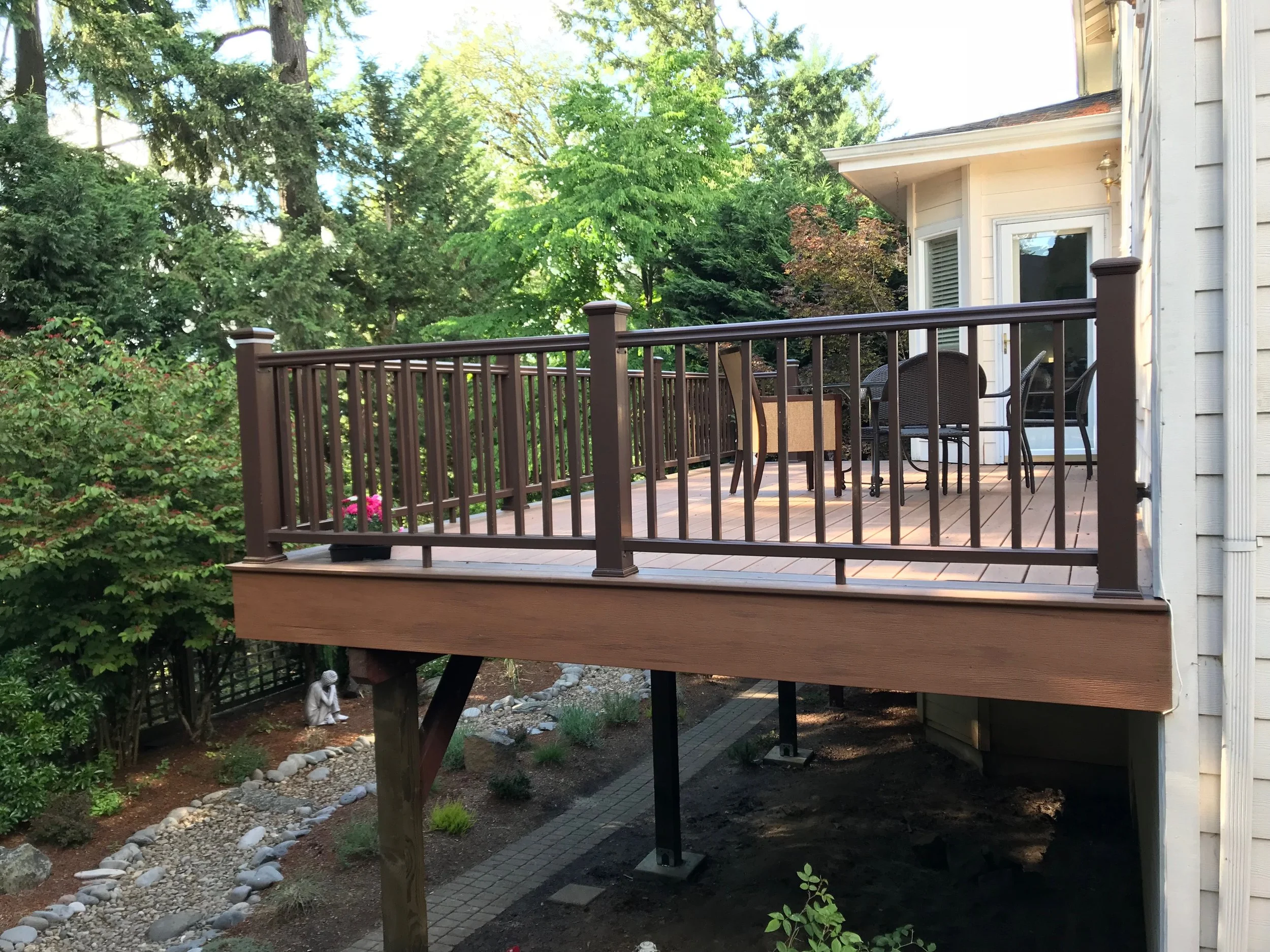 A wooden deck with a brown railing attached to a white house with a sliding glass door and window. There are chairs and a small table on the deck. Below the deck, there is a garden with rocks, small plants, and trees.