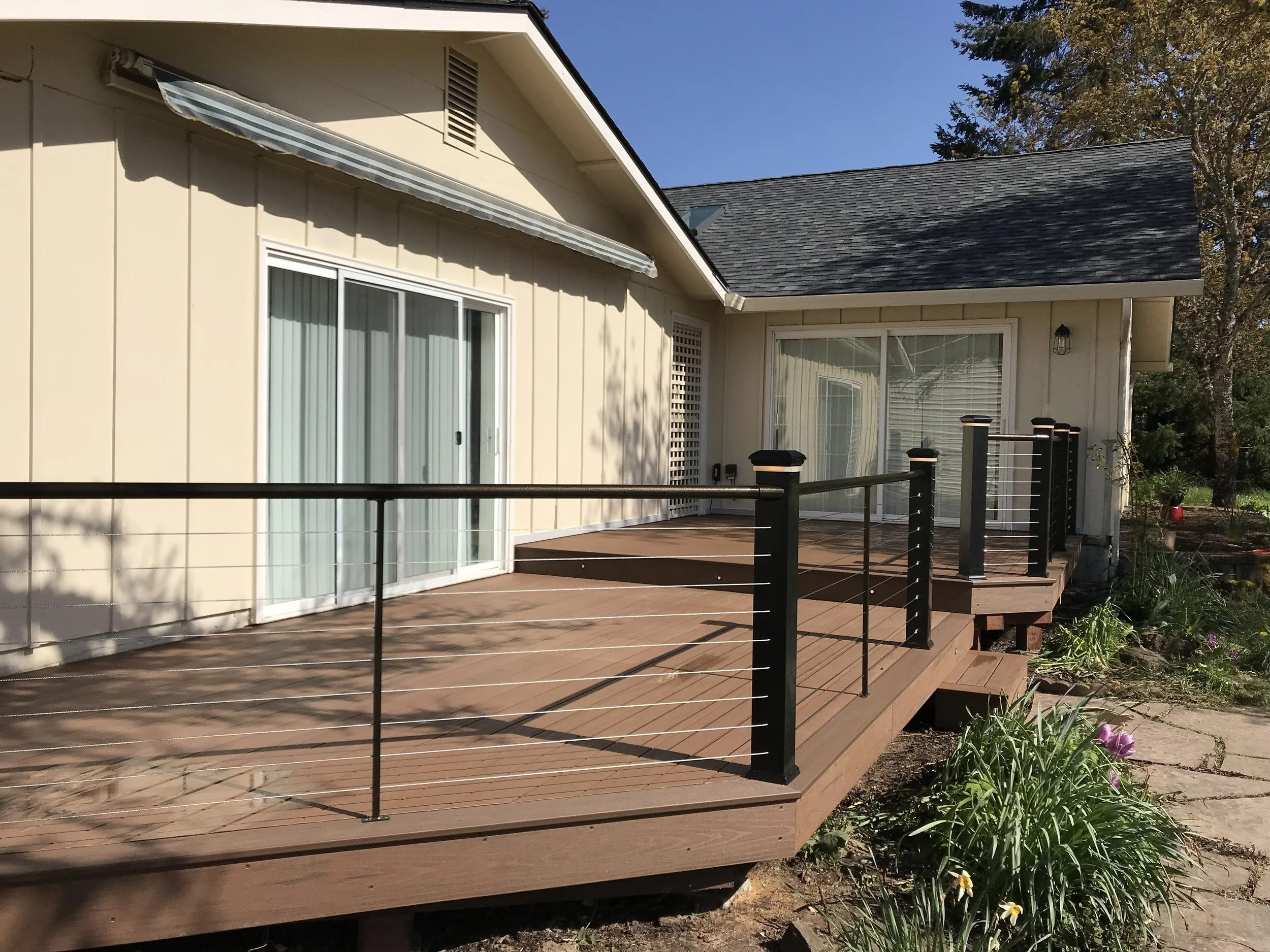 The image shows the exterior of a beige house with large sliding glass doors leading to a wooden deck with a black railing. The house has a gabled roof, a small vent near the roof’s peak, and a wall-mounted outdoor light fixture. There are trees and 