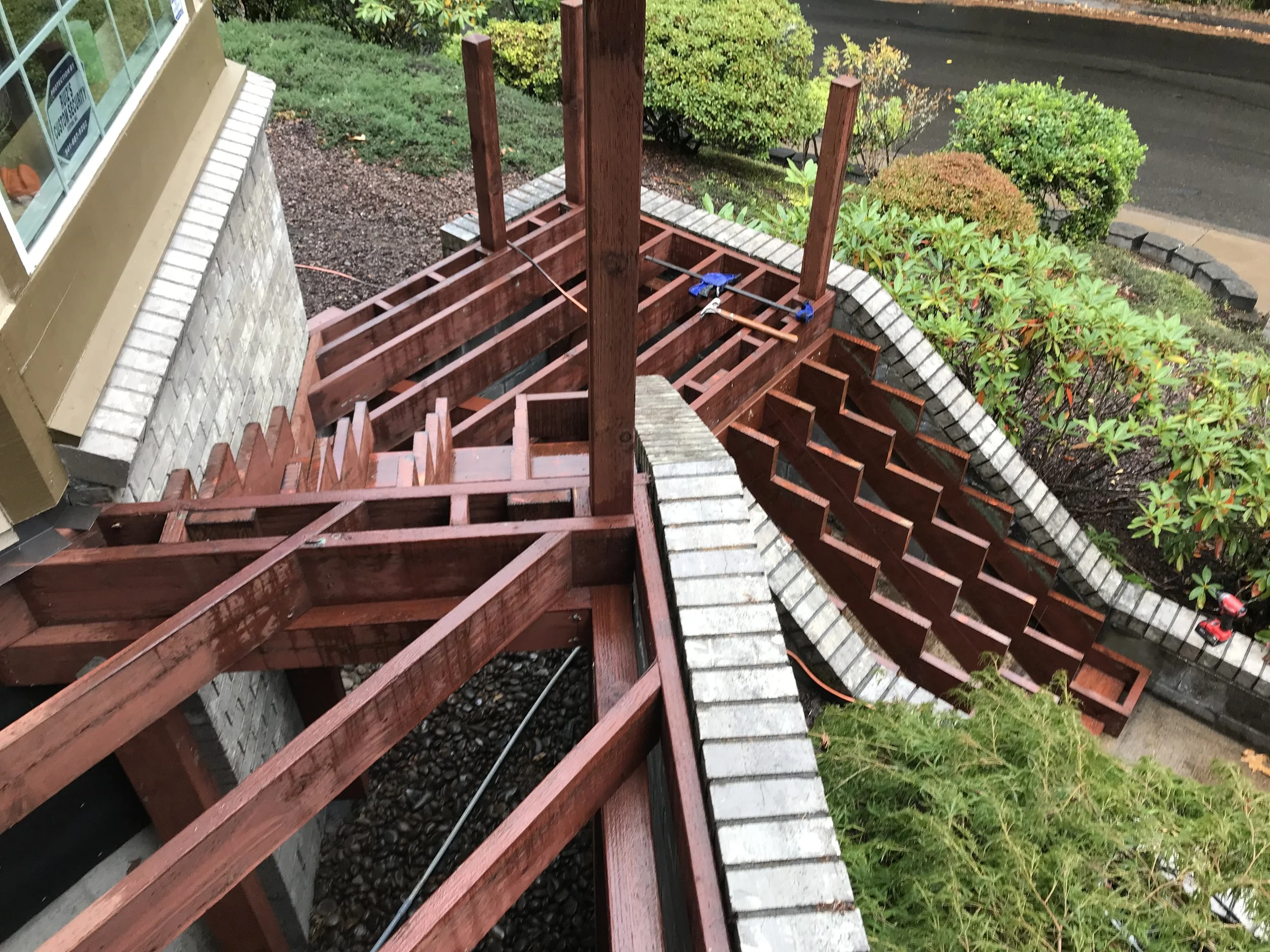 A wooden deck under construction with parts of the deck unfinished, showing the framework and support beams, next to a building with a window and a garden area with bushes and a sidewalk.