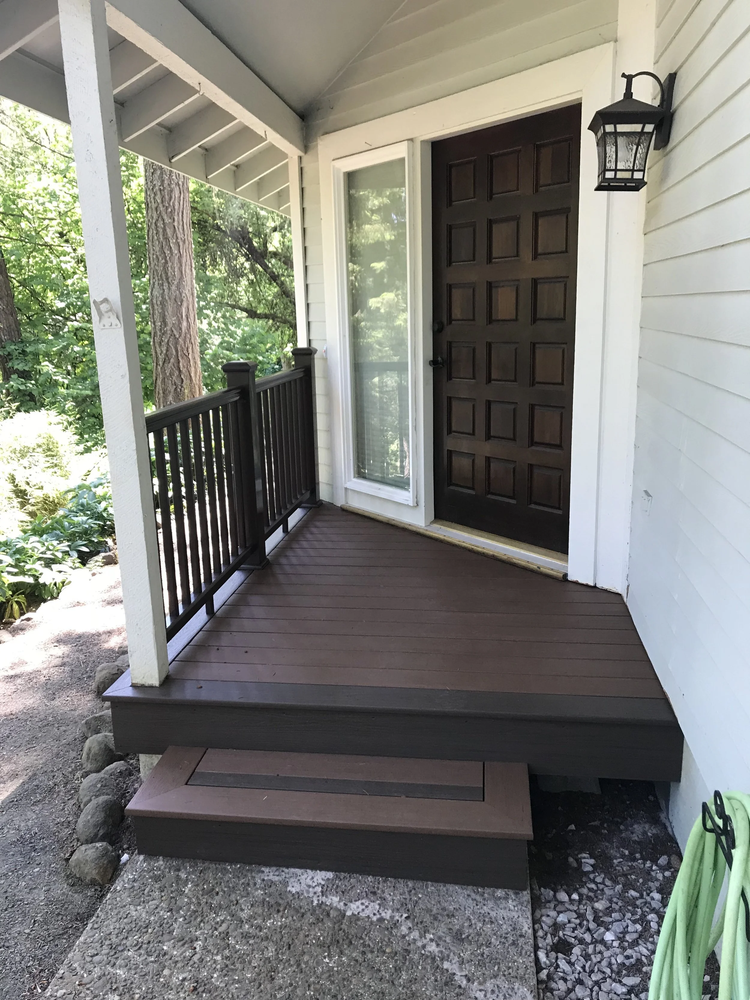 Front porch with dark brown wooden steps, black railing, white exterior wall, front door, and outdoor lantern light fixture.