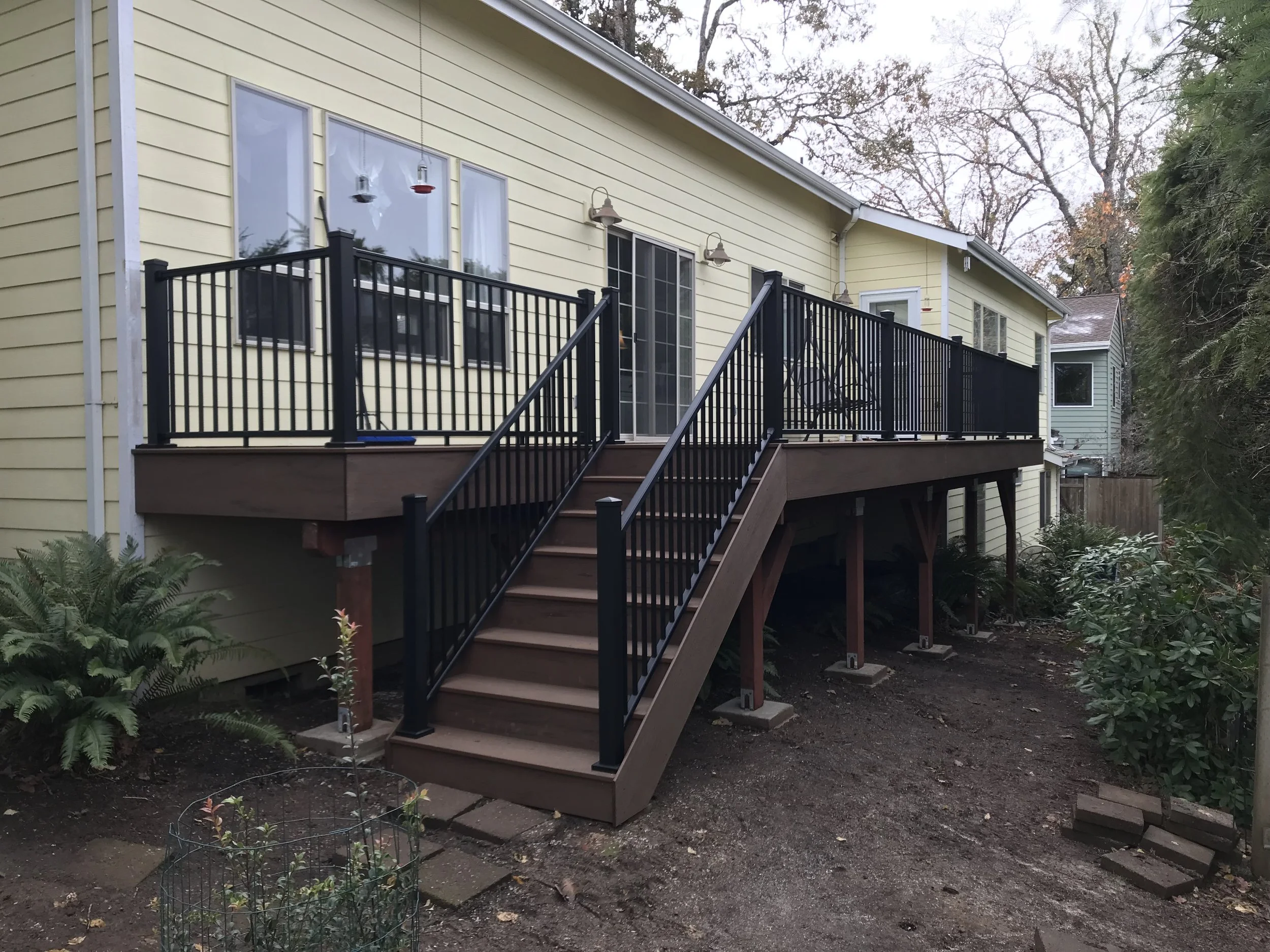 A yellow house with a black wooden deck and stairs in a backyard, surrounded by trees and plants.