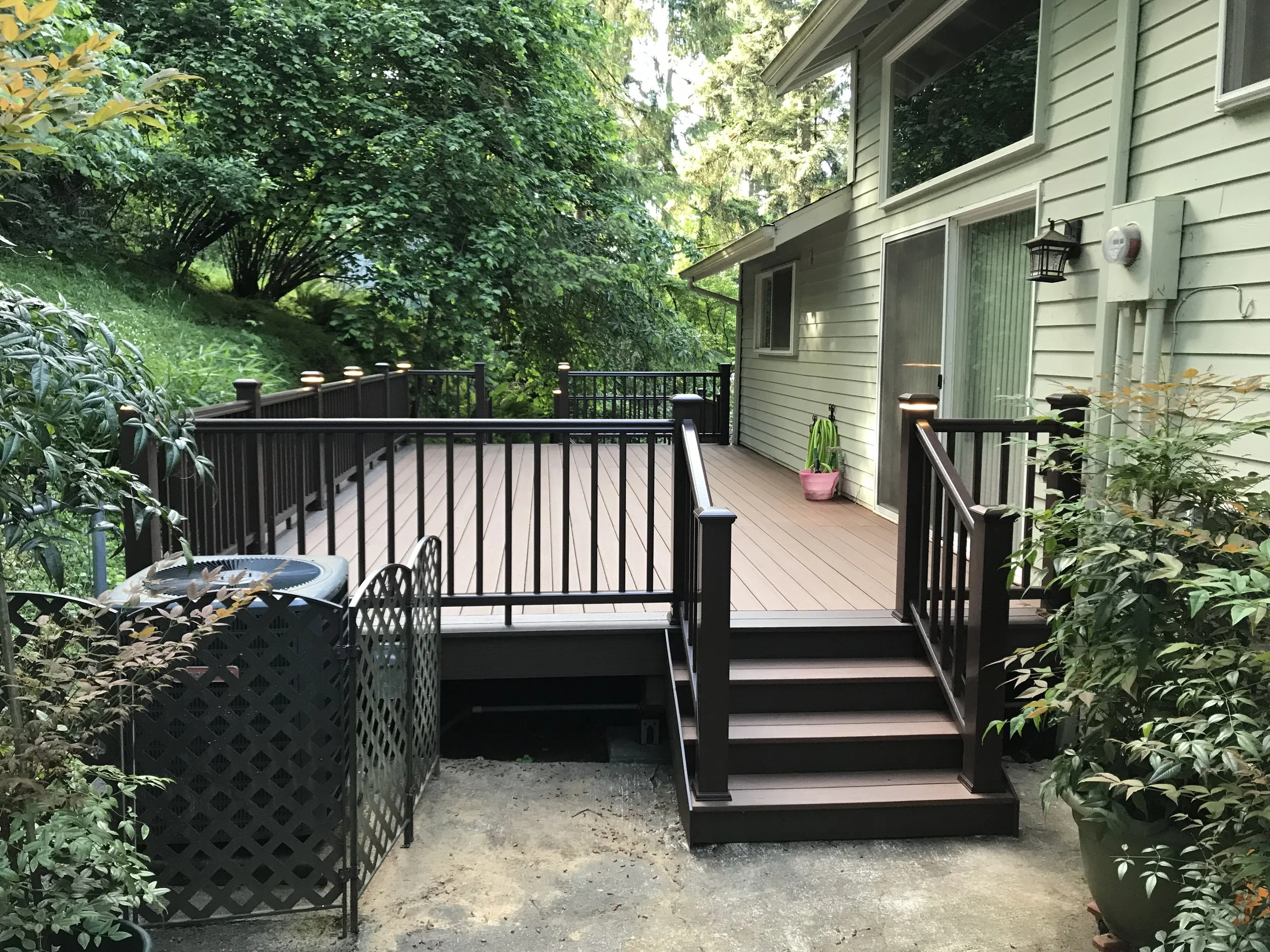 A home backyard with a wooden deck and black railing, surrounded by lush green trees and bushes, with a pink plant pot on the deck, and a house with light-colored siding and windows.