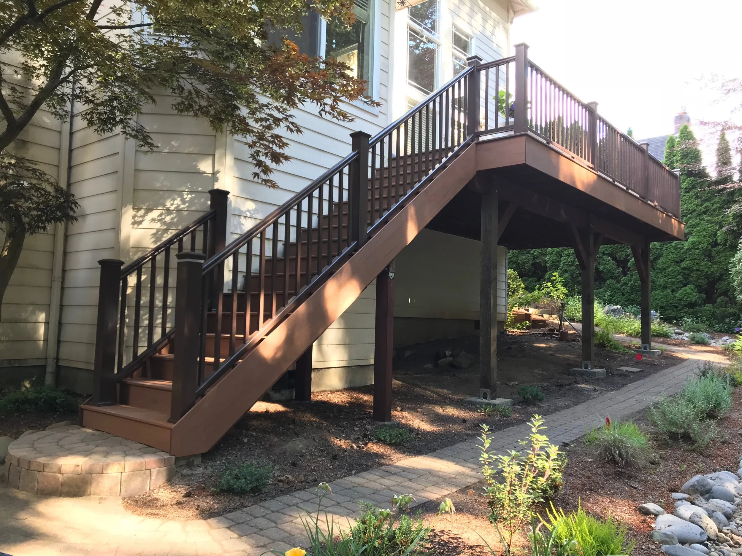 Backyard with a wooden deck with stairs, surrounded by trees and garden plants, next to a house with siding.