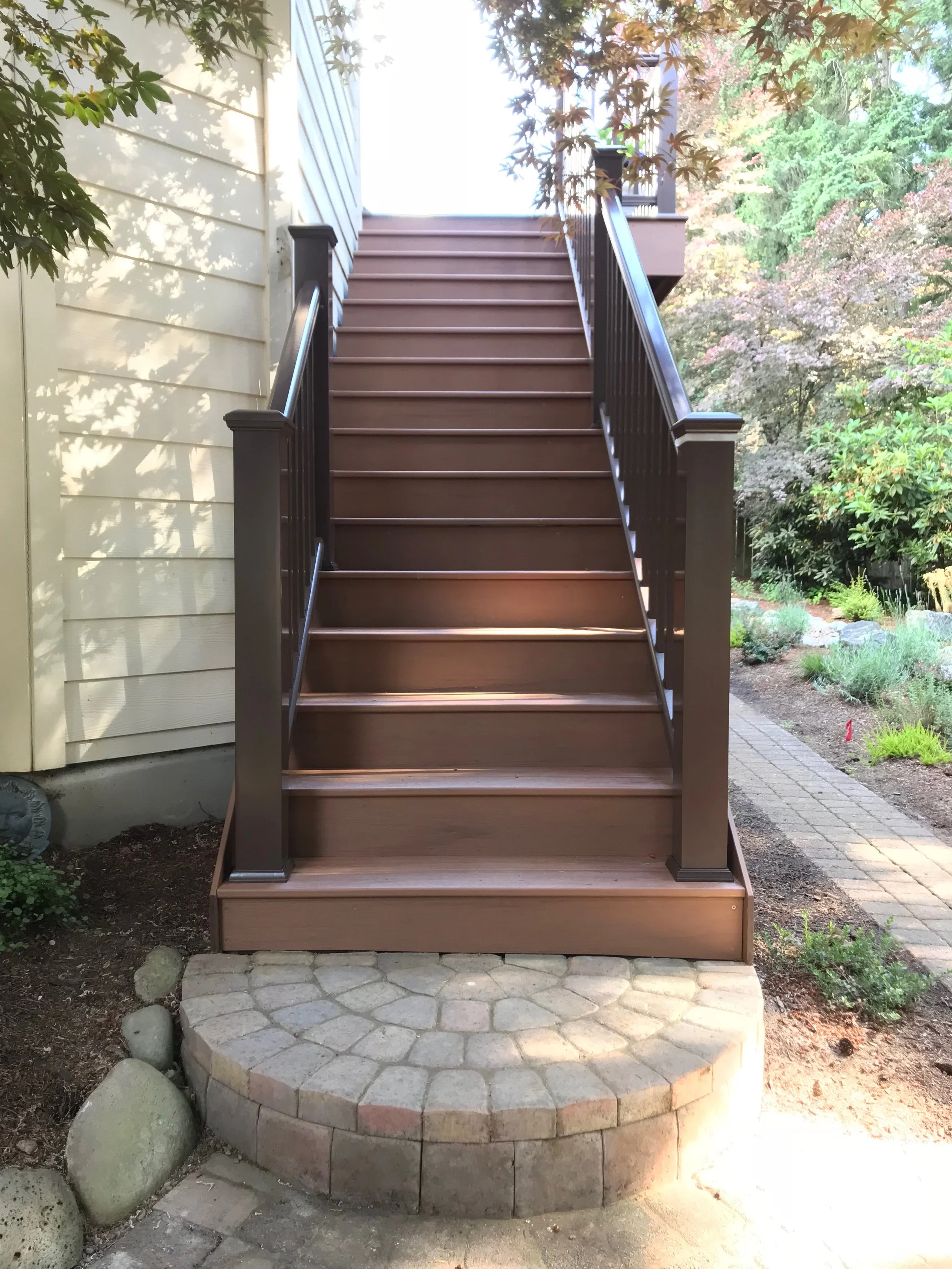 Wooden staircase with black railings leading up to a house, surrounded by trees and a stone-paved circular platform at the base.