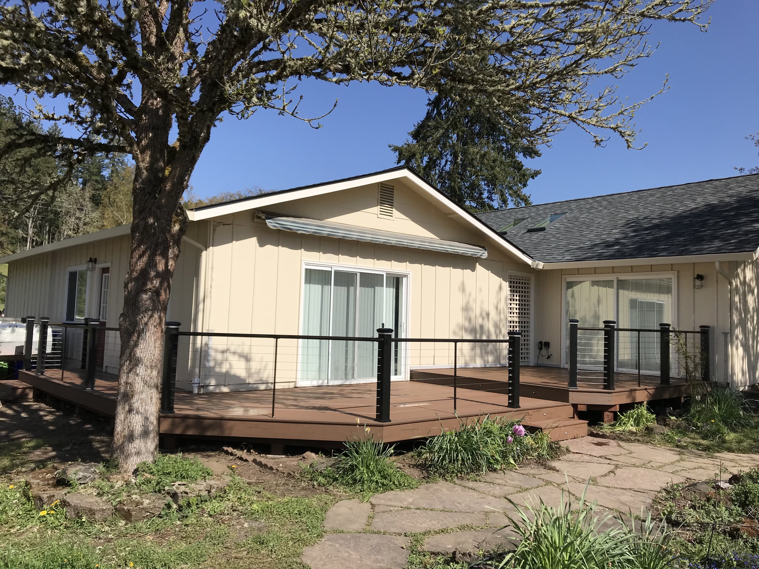 A backyard with a wooden deck, surrounded by green grass, plants, and flowers, with a beige house and large windows, a large tree with moss on the branches, and clear blue sky in the background.