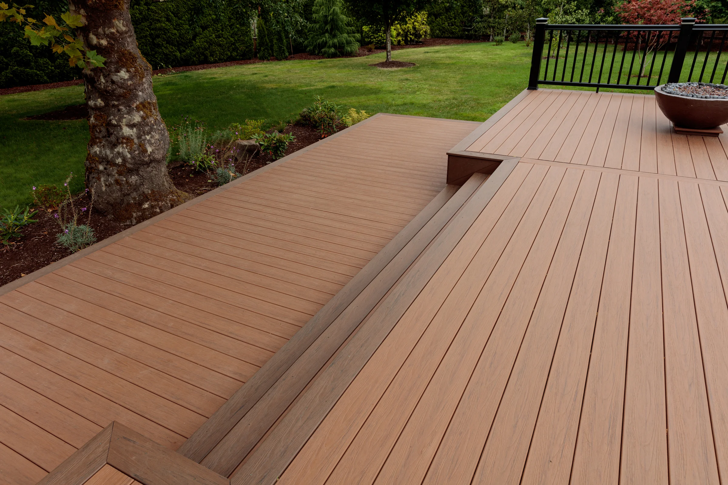 Backyard with a wooden deck area, a large tree on the left, green grass, and various trees and shrubs in the background. There is a black metal railing on one side of the deck and a large decorative pot filled with rocks.