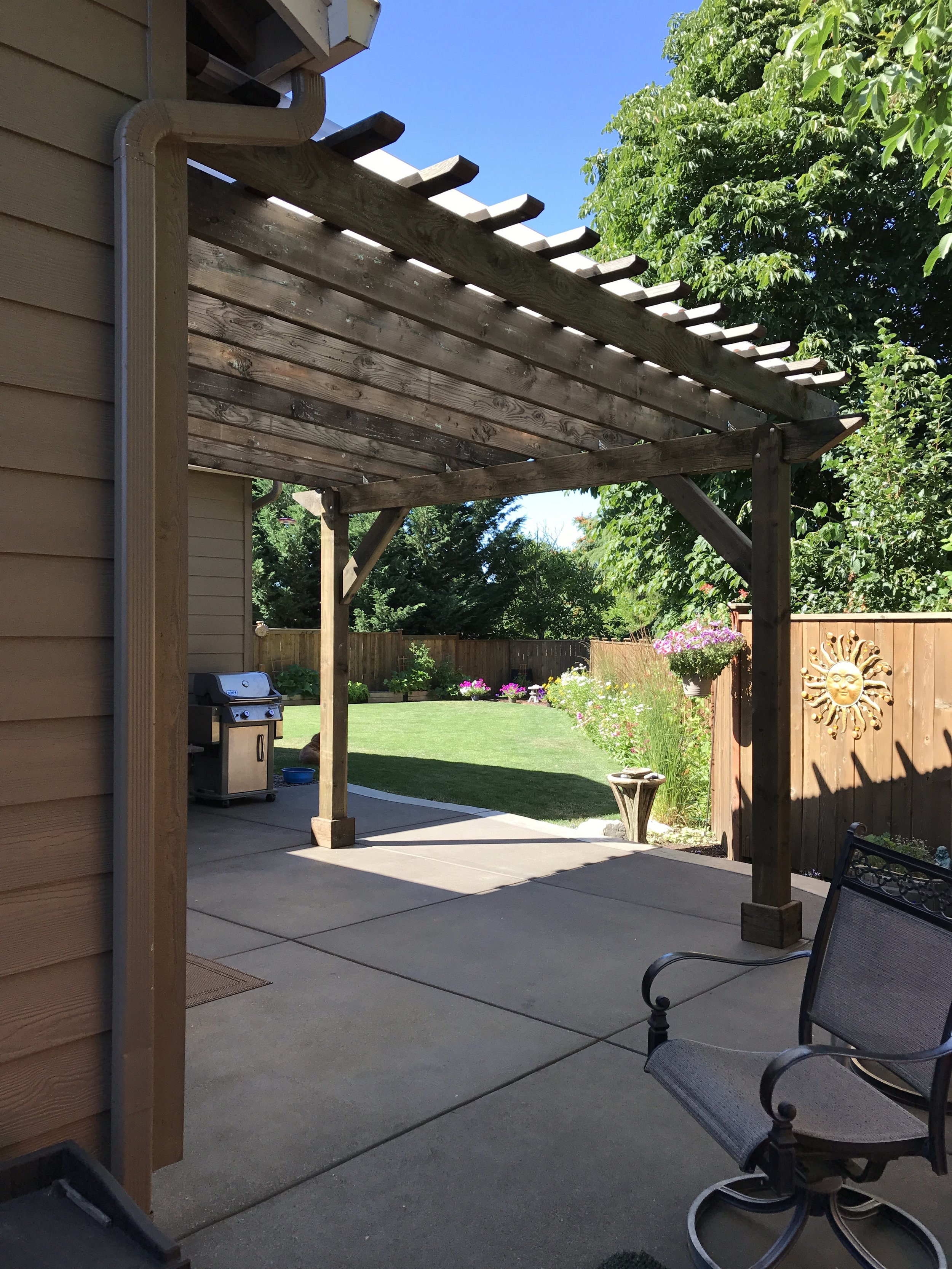 View of a backyard patio with a wooden pergola, outdoor chairs, a barbecue grill, a garden with colorful flowers, lush green trees, and a blue sky.
