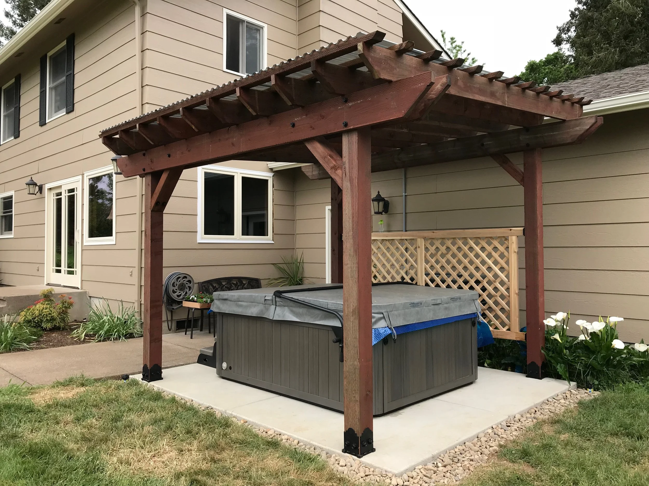 A backyard patio with a beige house in the background. There is a wooden pergola structure with a hot tub underneath it. The area is landscaped with grass, small plants, and a stone border.