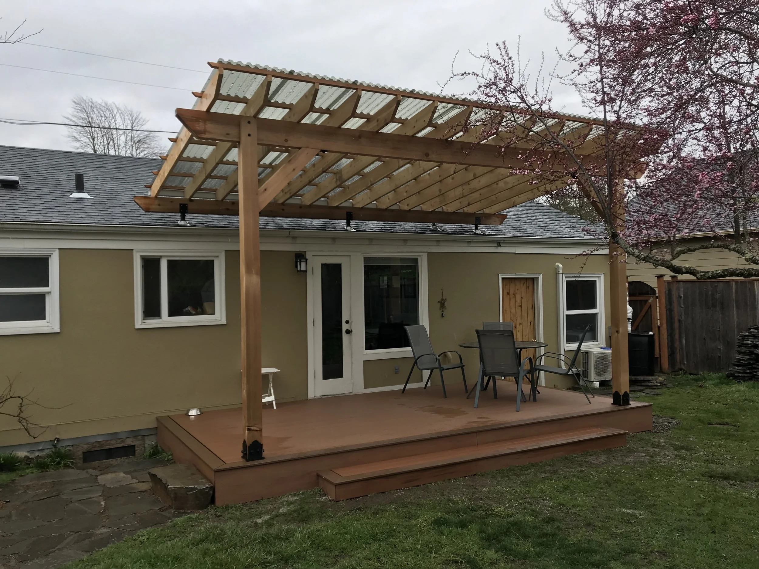 A wooden patio cover being built over a backyard deck, attached to a house with beige siding and white-framed windows, with outdoor furniture and a flowering tree nearby.
