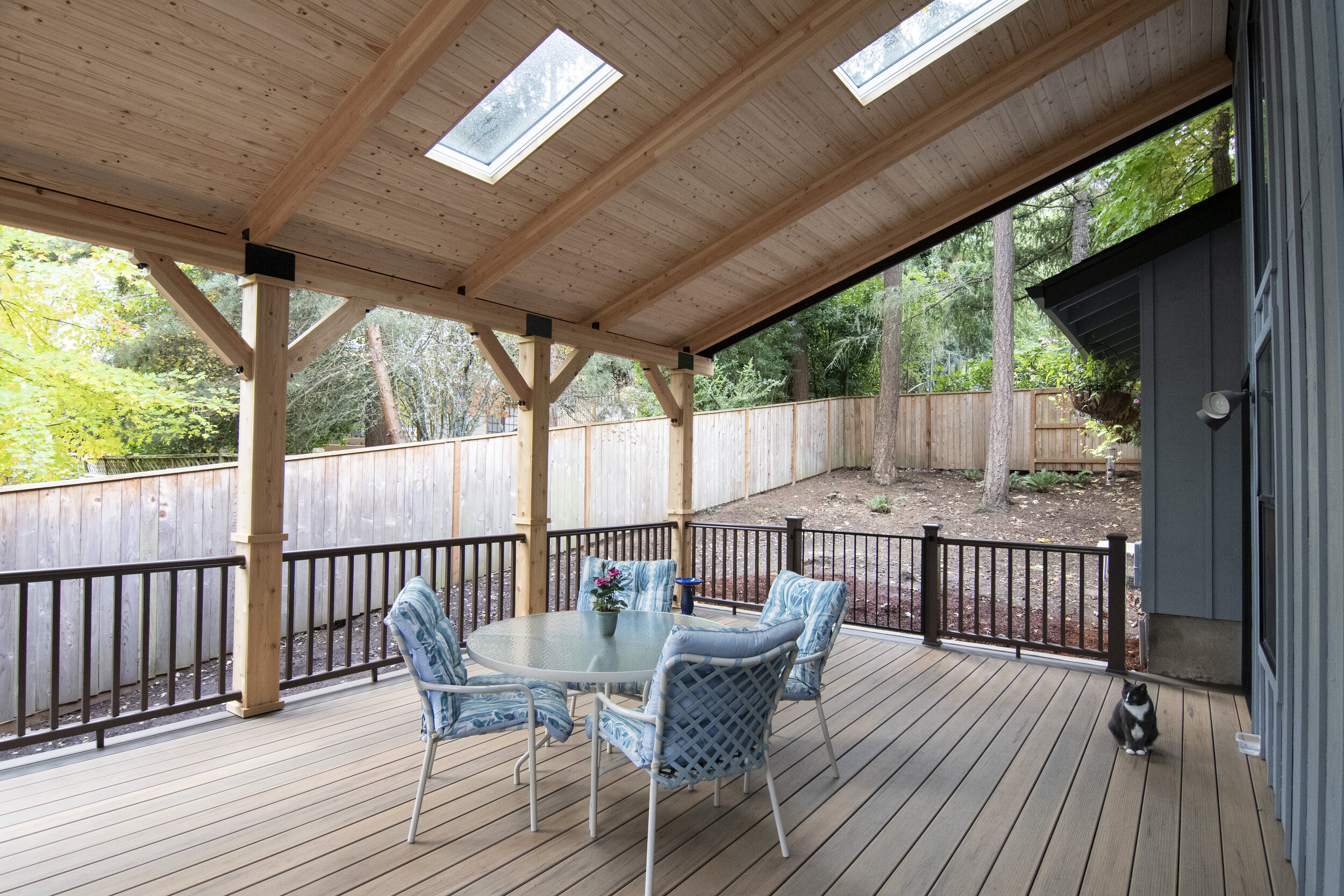 Covered outdoor patio area with a round glass table, four chairs with blue and white striped cushions, a small potted plant, and a black and white cat sitting at the edge near a blue house wall. The patio has a wooden ceiling with skylights, wooden s