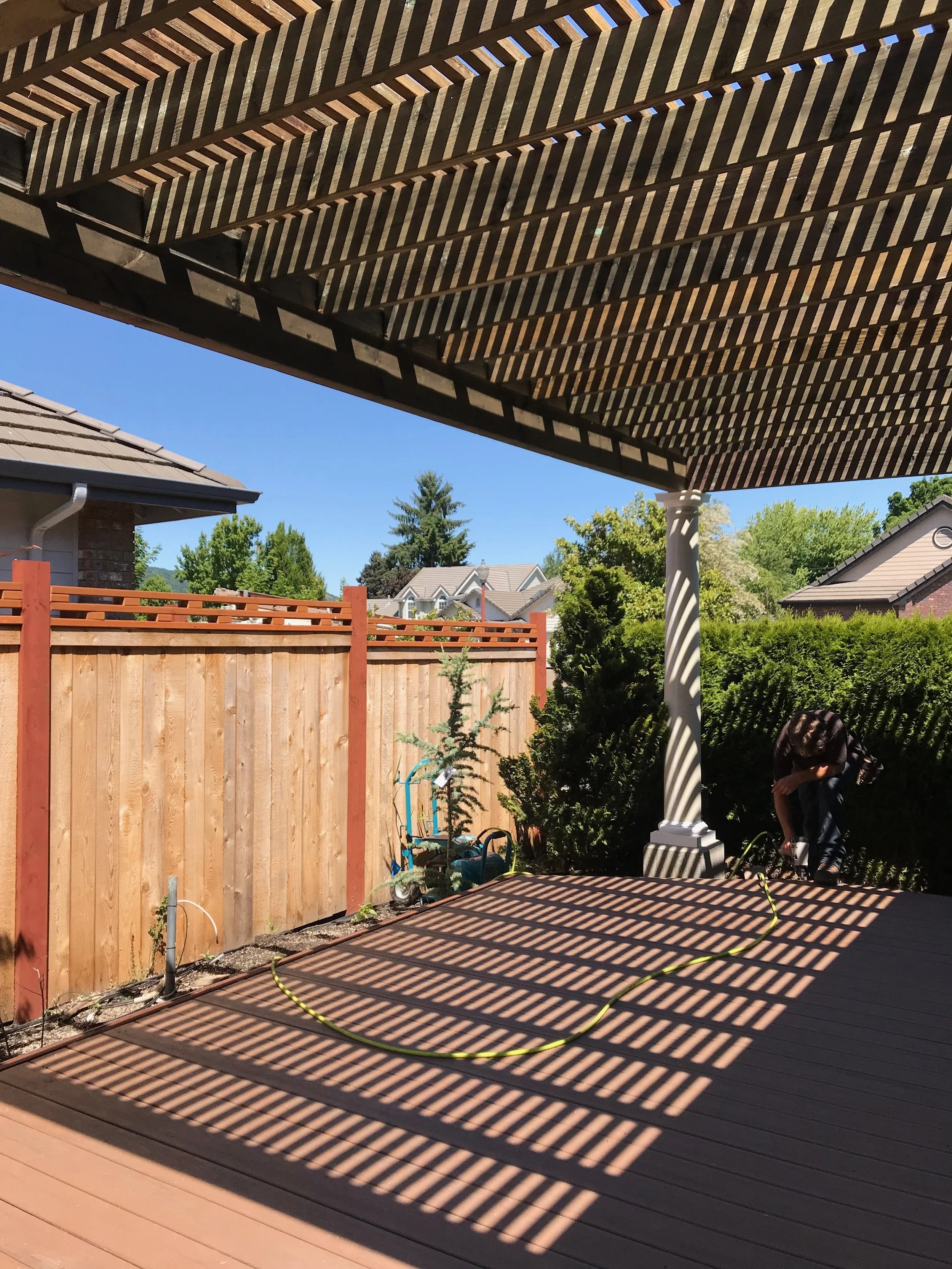 View of a backyard deck with sunlight casting striped shadows through a slatted pergola. A wooden fence runs along the yard, with some garden tools and a small tree visible. In the background, trees and houses are seen under a clear blue sky.
