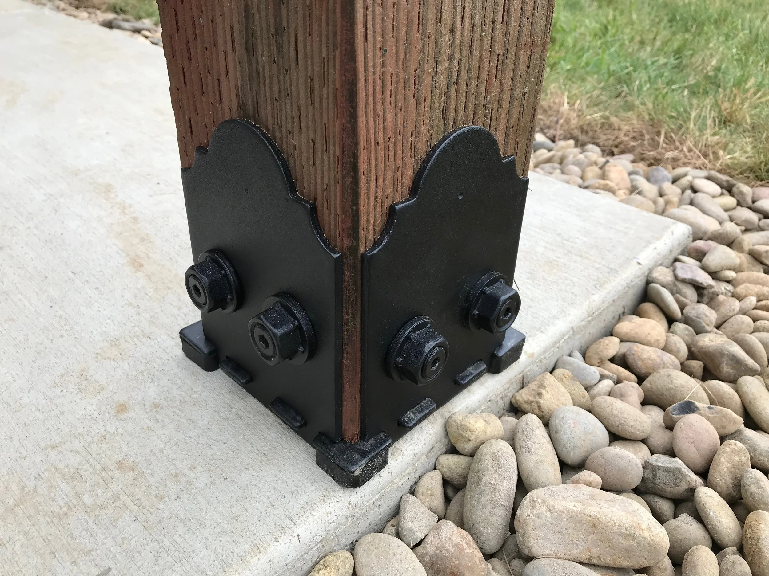 A wooden post secured in a black metal post base attached to a concrete slab, with smooth river rocks surrounding it on the ground.