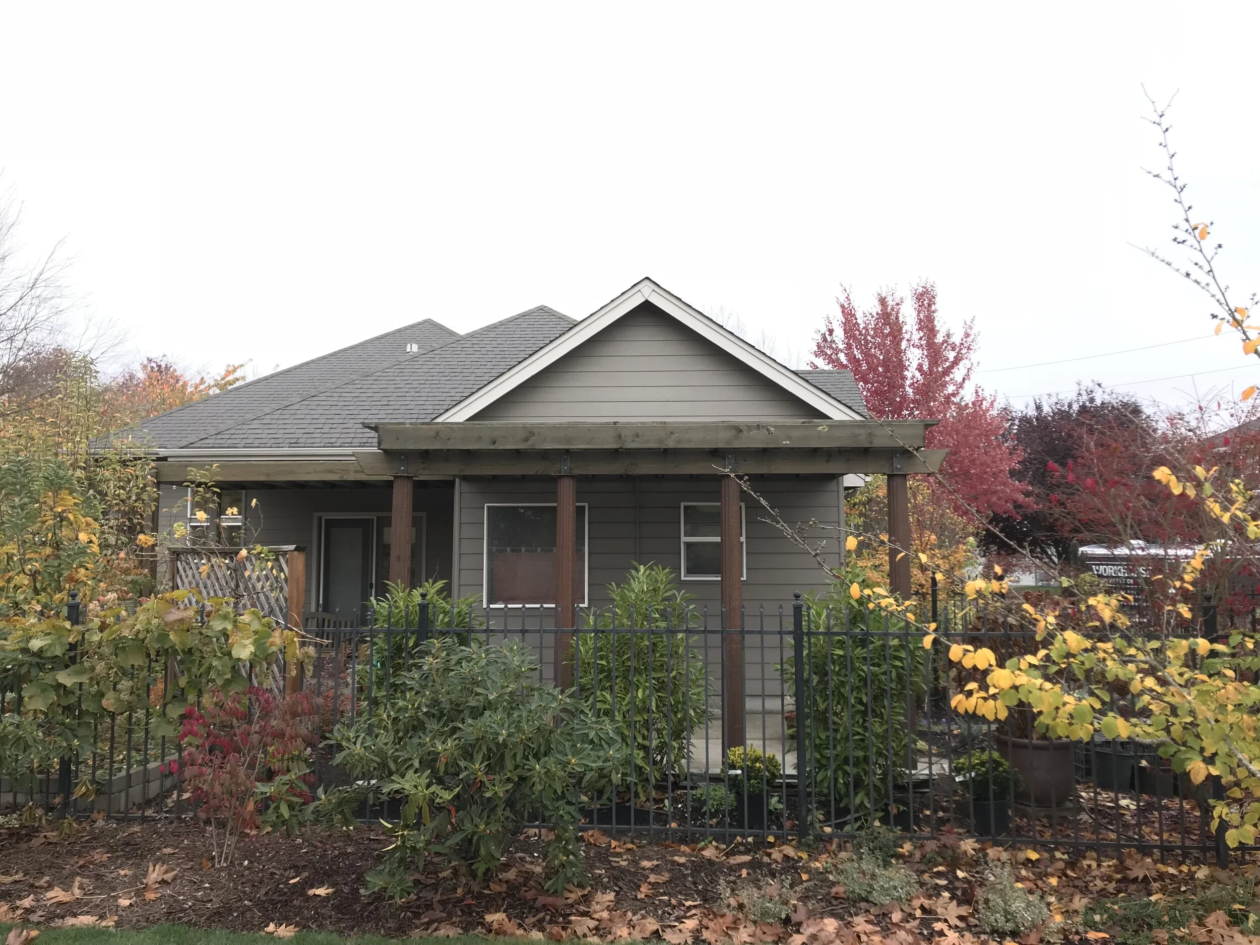 A house with gray siding and a gray shingled roof, surrounded by autumn trees and bushes with yellow, red, and orange leaves, a garden, and a black metal fence in the foreground.