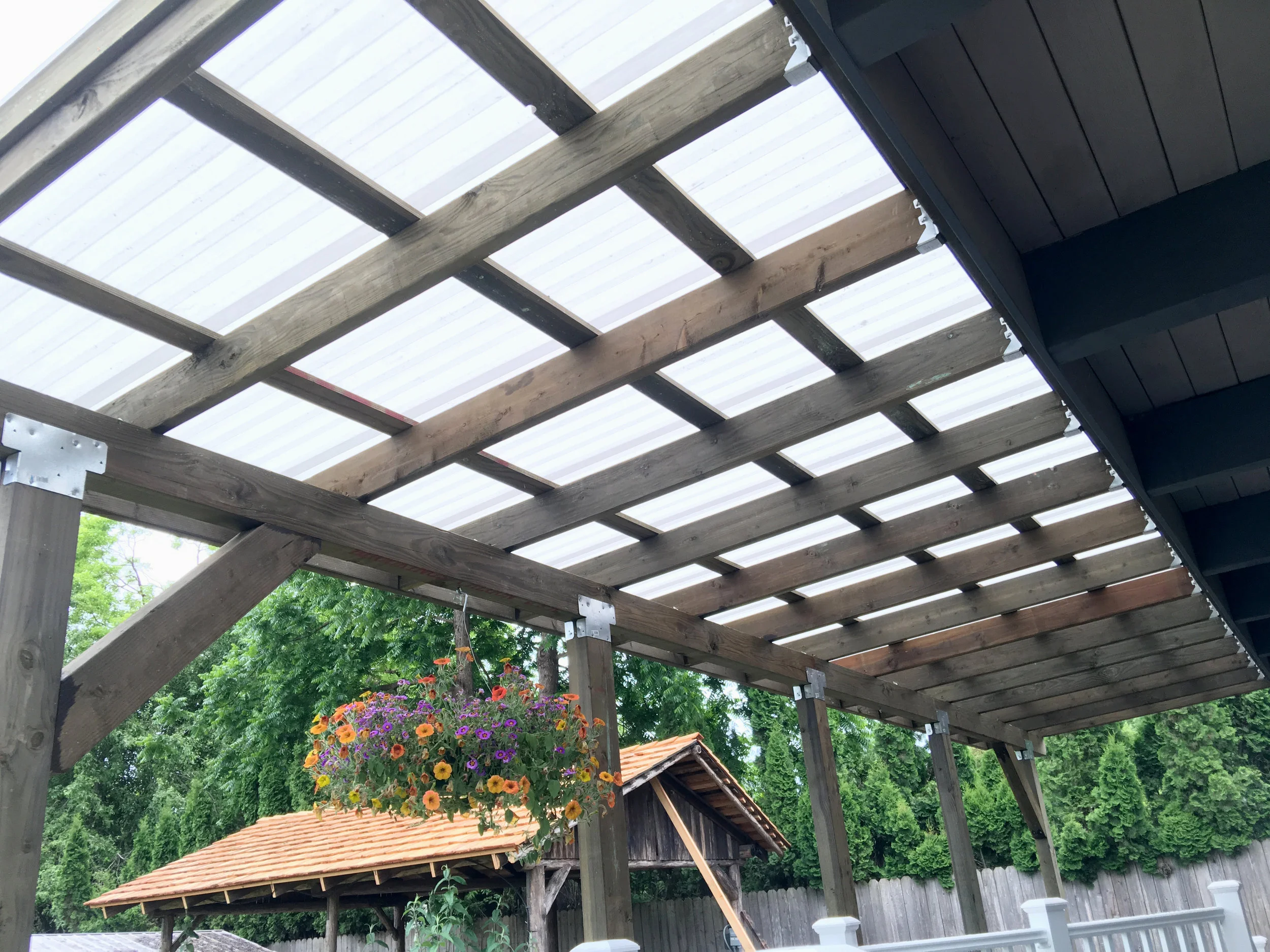 Photo of a wooden pergola with beams and a translucent roof, blue siding on the structure's side, background trees, a hanging flower basket with purple, orange, and yellow flowers, a shed with a tiled roof, and a wooden fence.