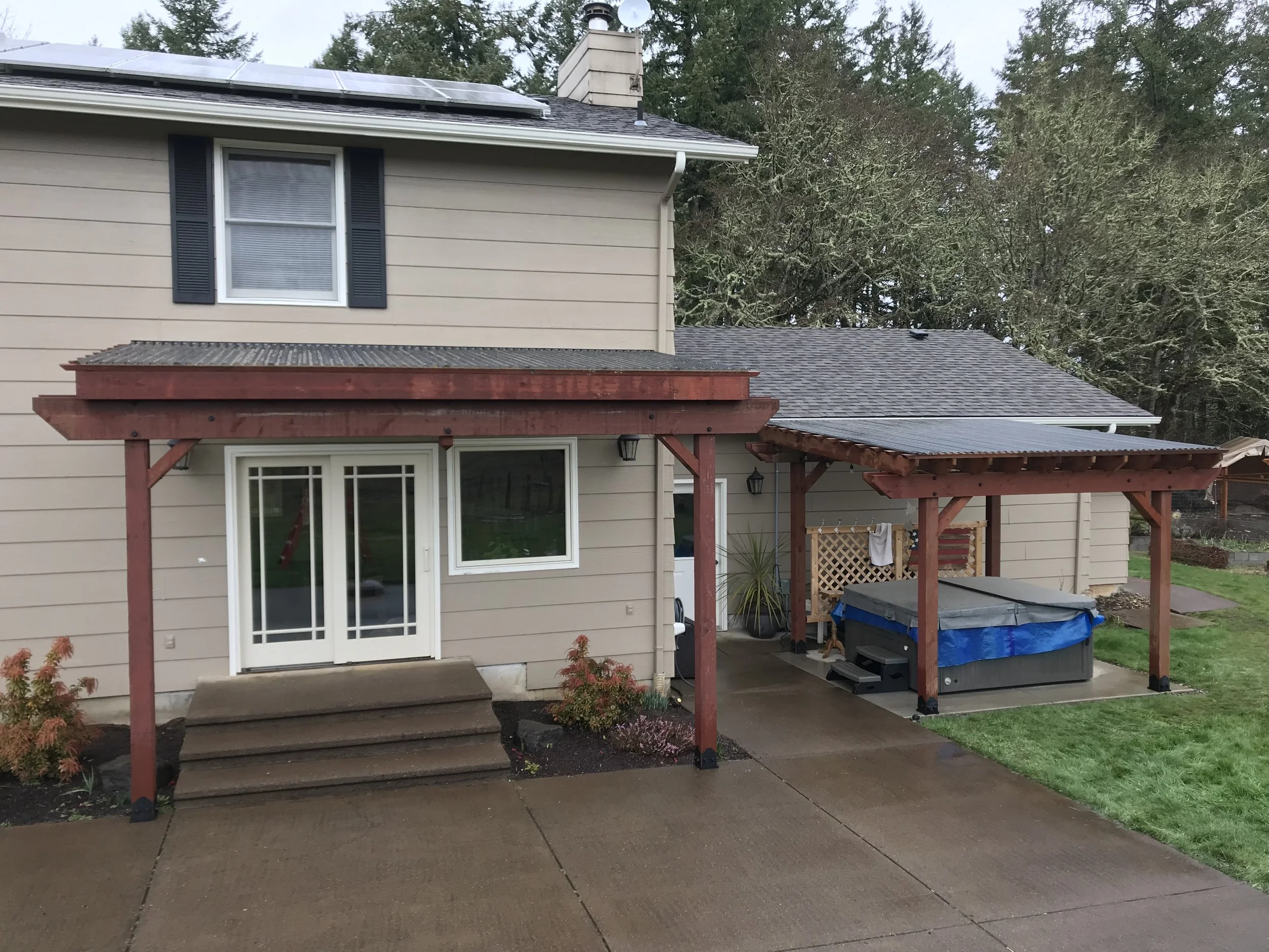 View of the back of a beige house with black window shutters, a sliding door, a wooden pergola, and a patio area with gardening supplies, surrounded by trees and a cloudy sky.