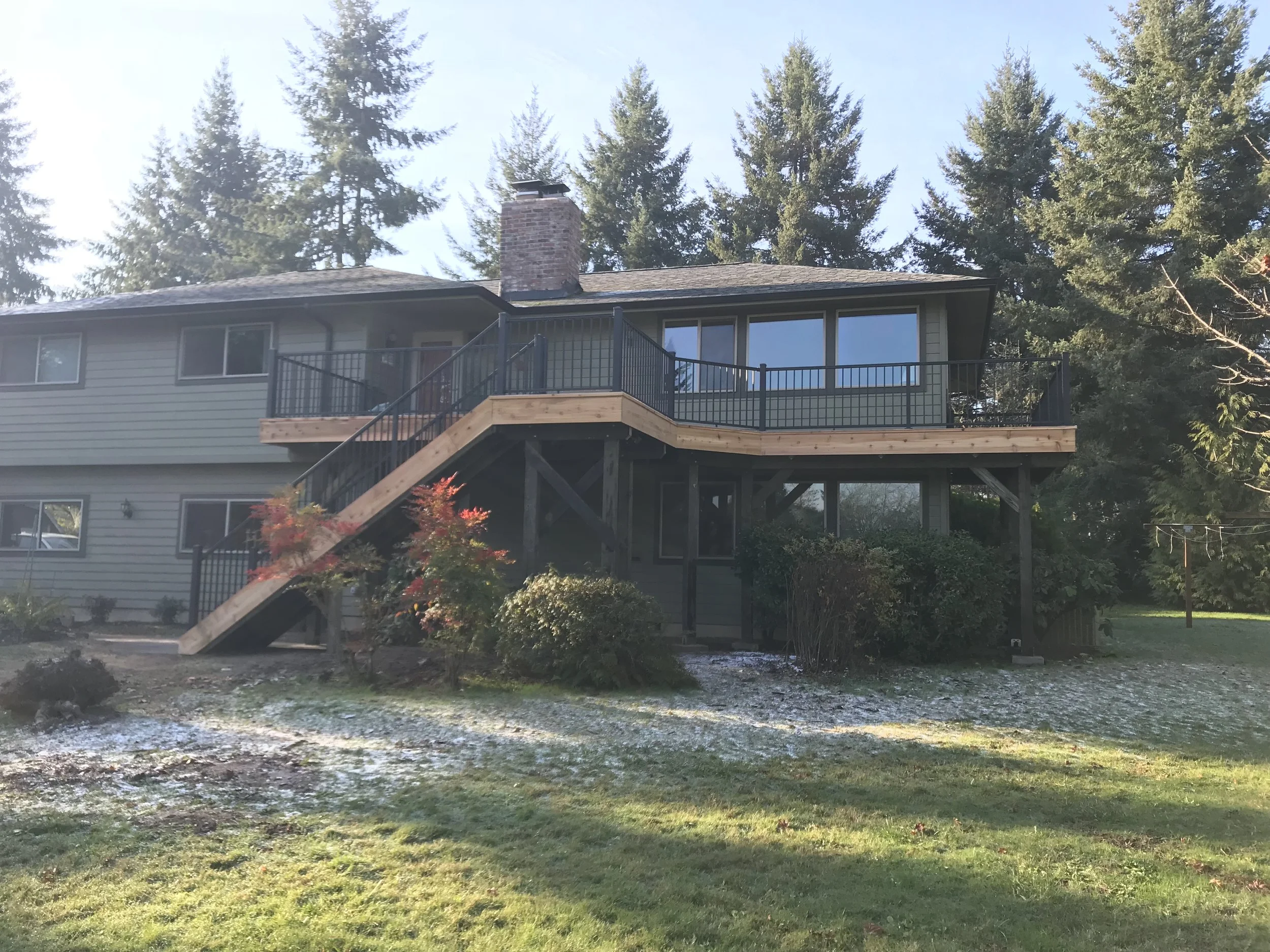 A two-story house with gray siding, large front windows, a brick chimney, and a wooden deck with black railings in a backyard surrounded by tall evergreen trees.
