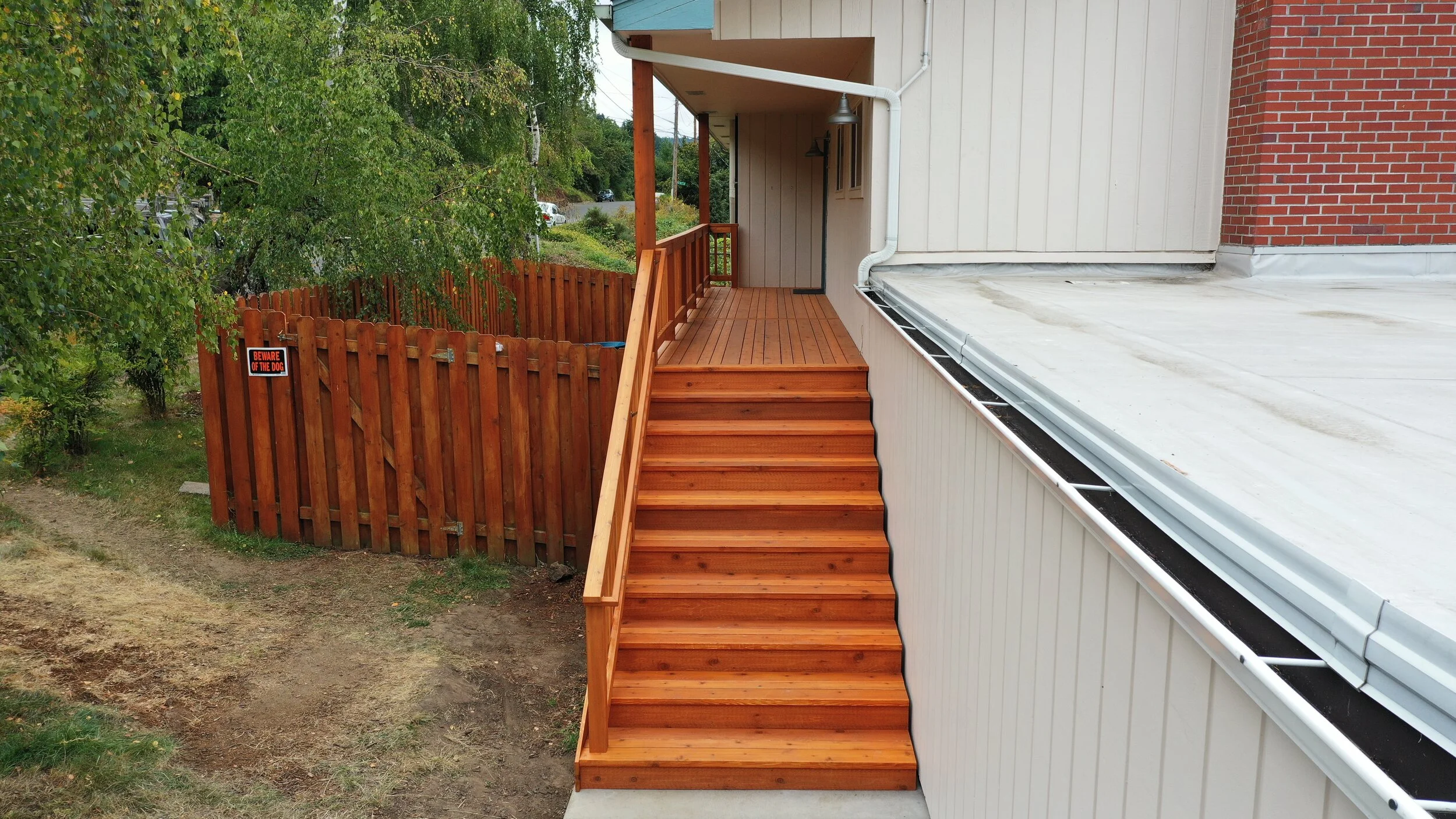 Wooden stairs leading up to a porch with railing, next to a white exterior wall of a building, and a fence with a warning sign about a dog.