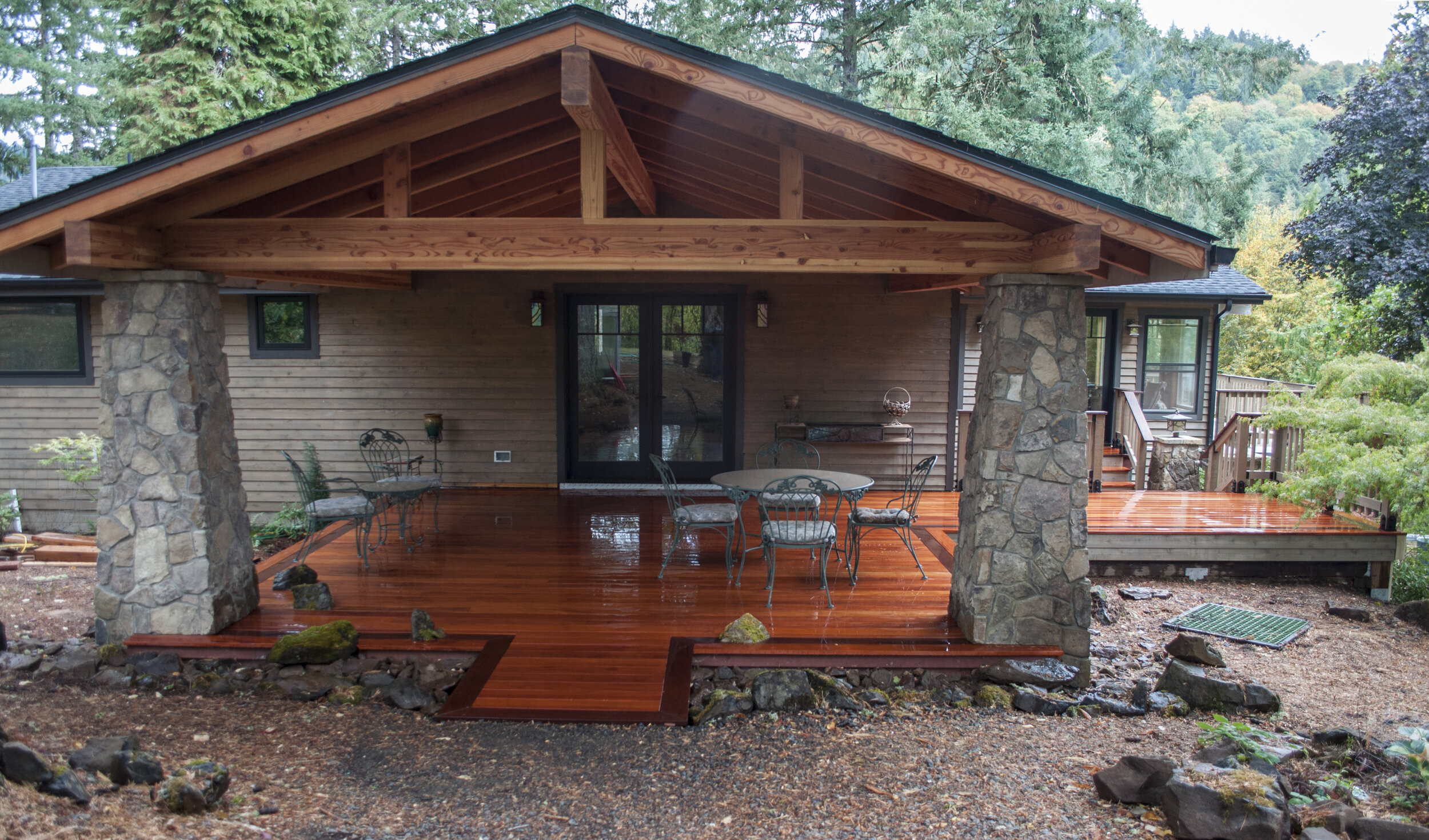 Back view of a house with a wooden deck, stone pillars, and large windows, surrounded by trees.