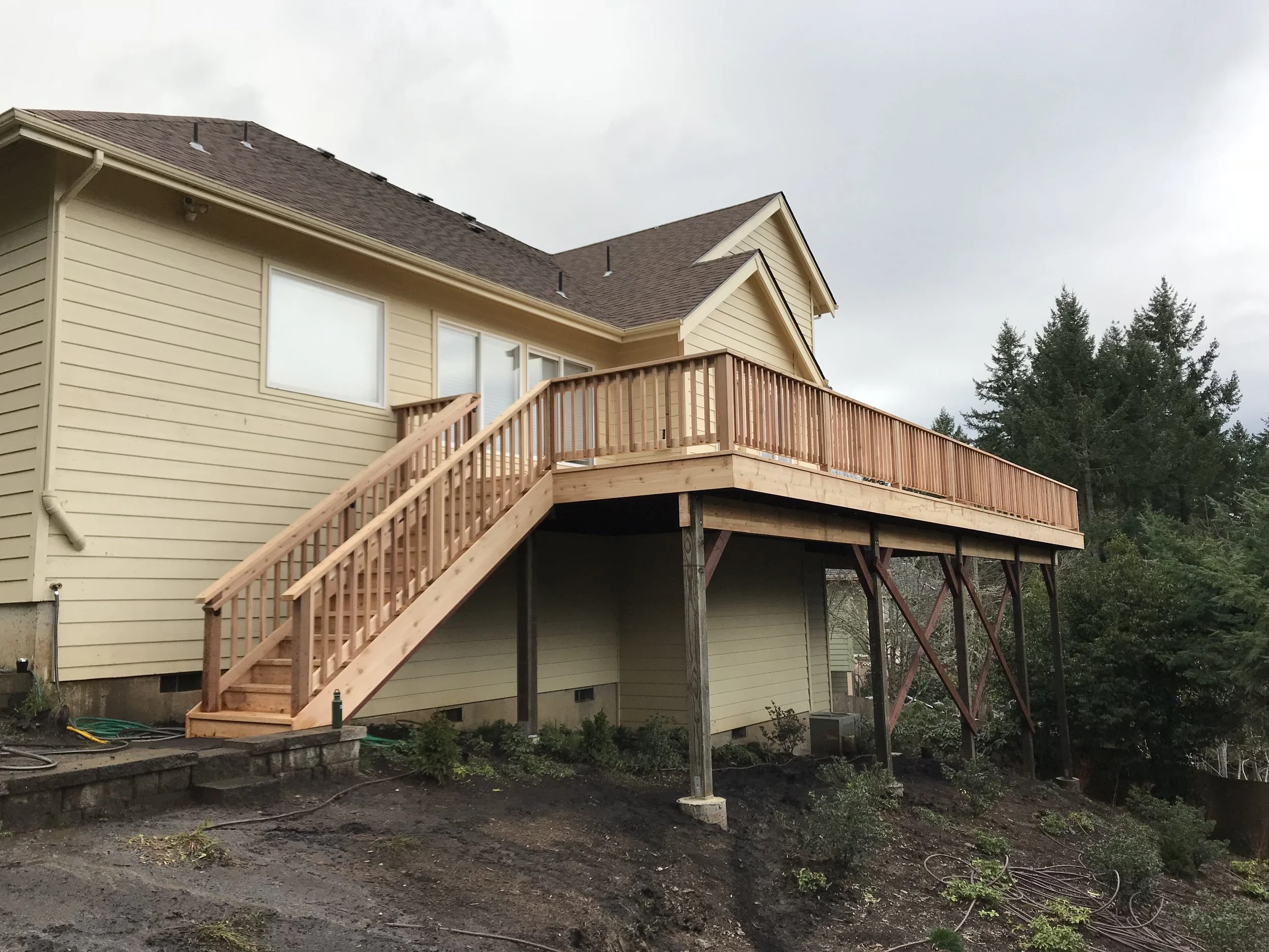A two-story house with beige siding, a wooden deck with stairs, and a sloped roof, situated in a yard with trees in the background.