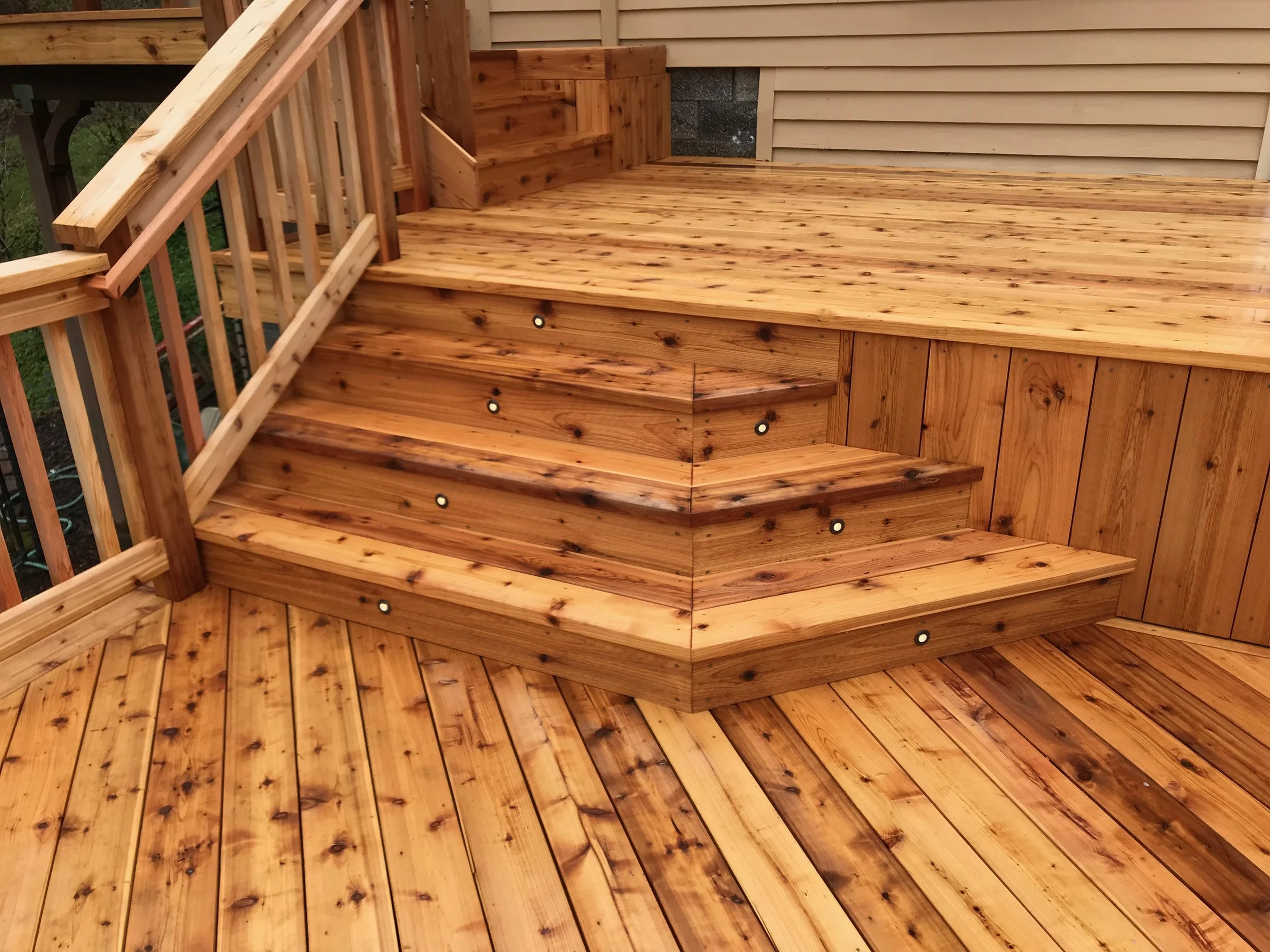 Newly constructed wooden deck with stairs, built using natural wood planks, attached to the side of a house with beige siding, featuring a small set of steps and a wooden railing.
