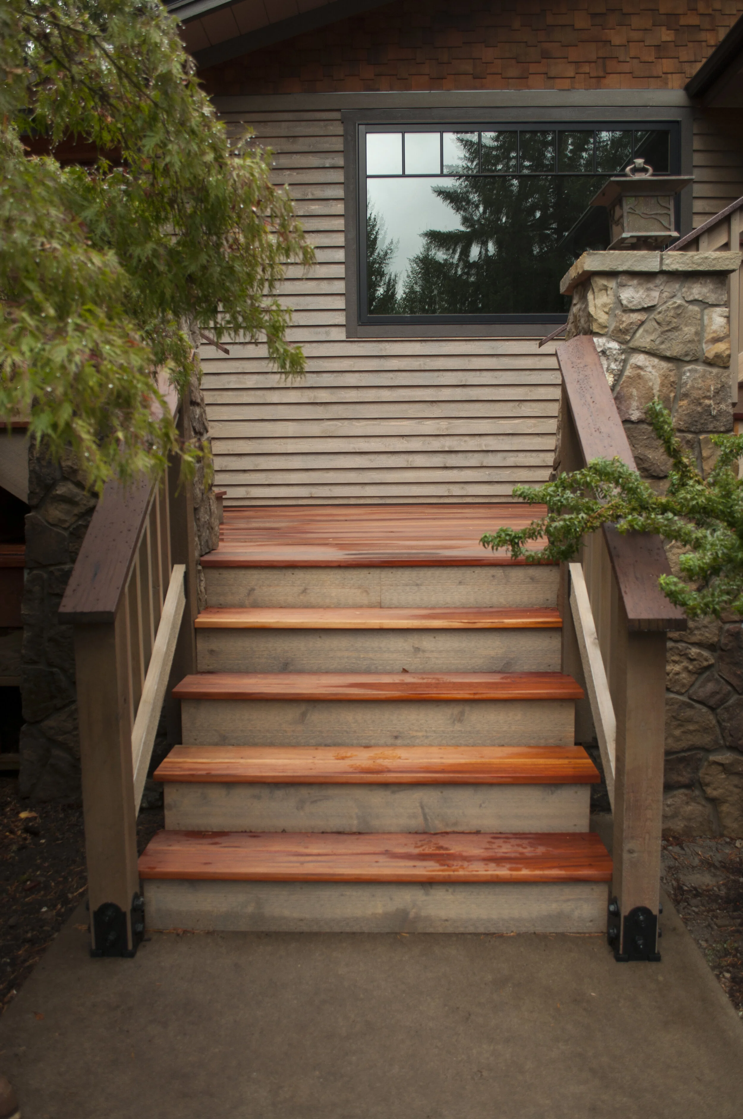 Wooden stairs leading up to a house with beige horizontal siding, a large black-framed window reflecting trees, a stone pillar on the right, and green trees around the stairs.