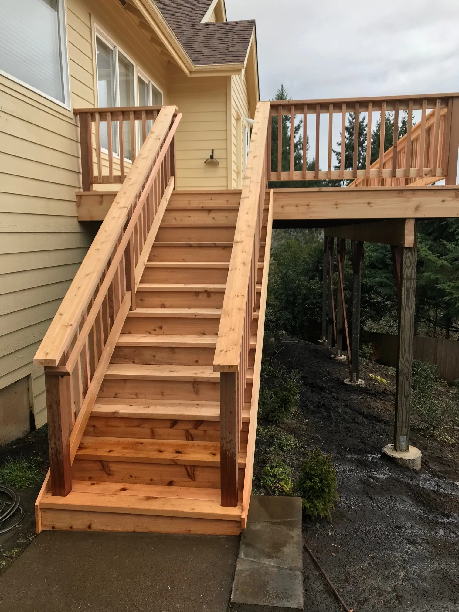 Newly built wooden deck with stairs leading up to it attached to a yellow house with beige siding. The deck has wooden railings and supports, and there are trees and cloudy sky in the background.