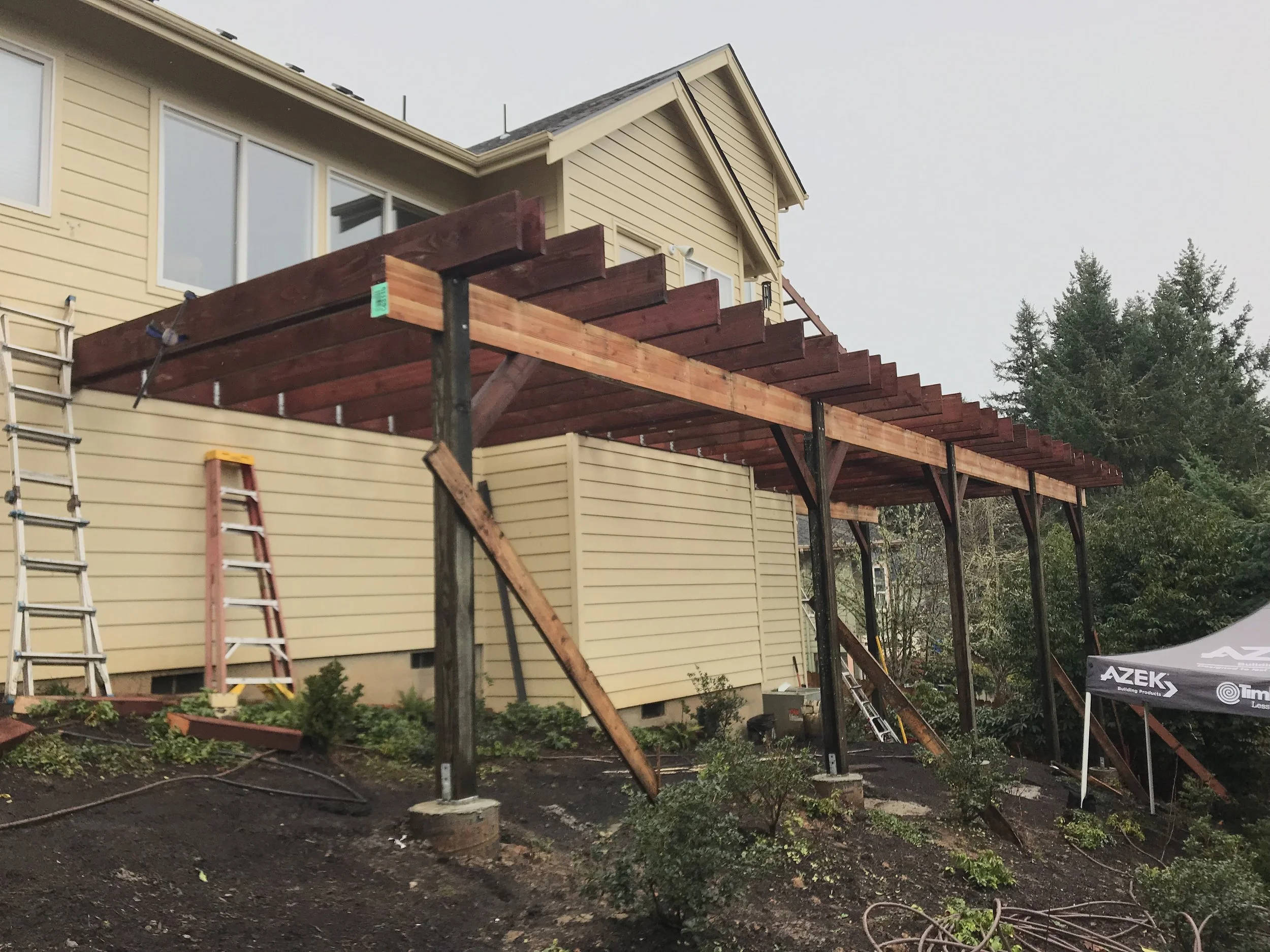 A house under construction with a large wooden deck framework built on support beams, scaffolding, ladders, and construction equipment in the yard.