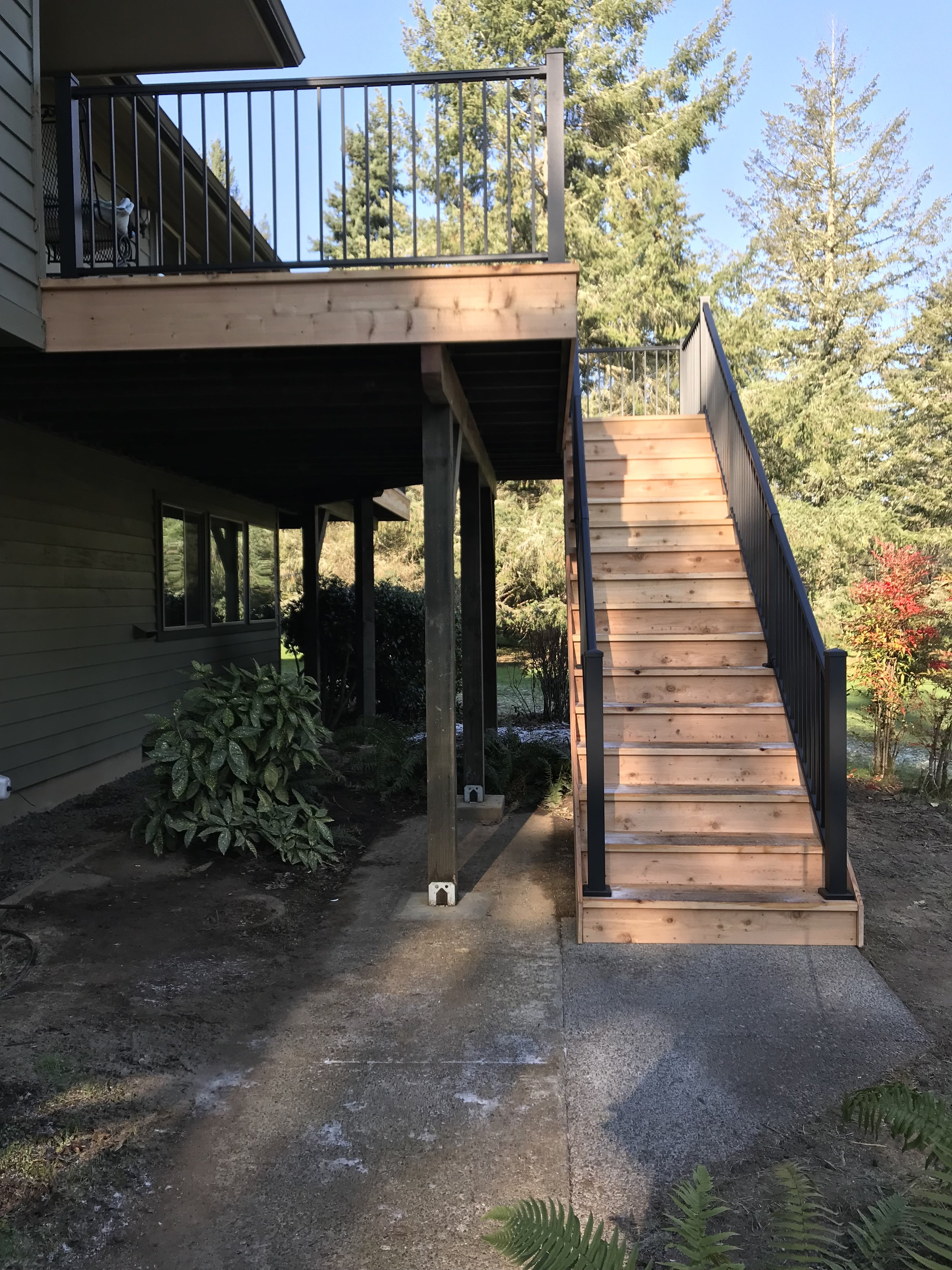 Wooden staircase with black metal railing leading to a house's second floor outdoor deck, with trees and blue sky in the background.