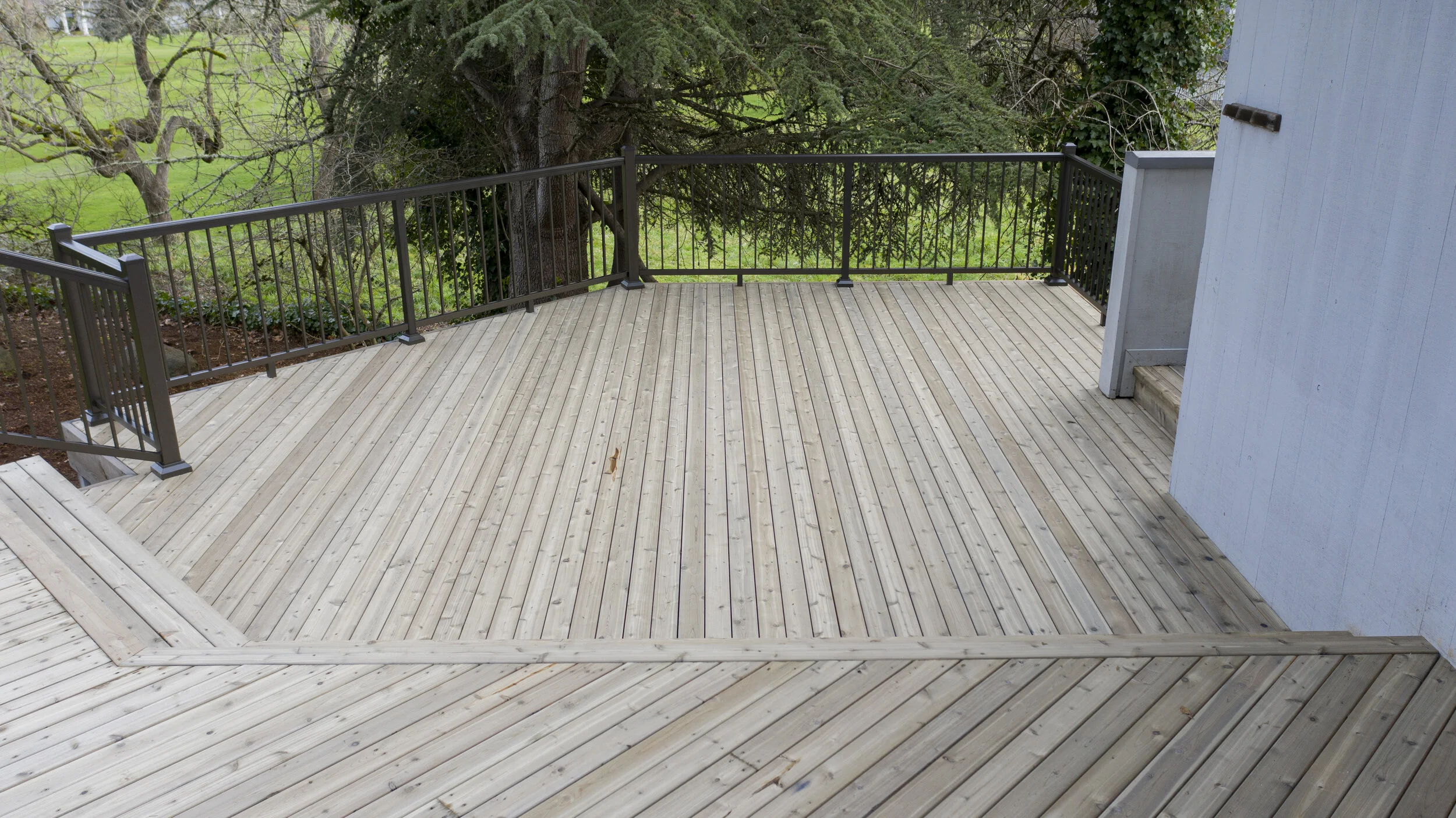 Wooden deck with black metal railing next to a house, overlooking a grassy yard with trees.