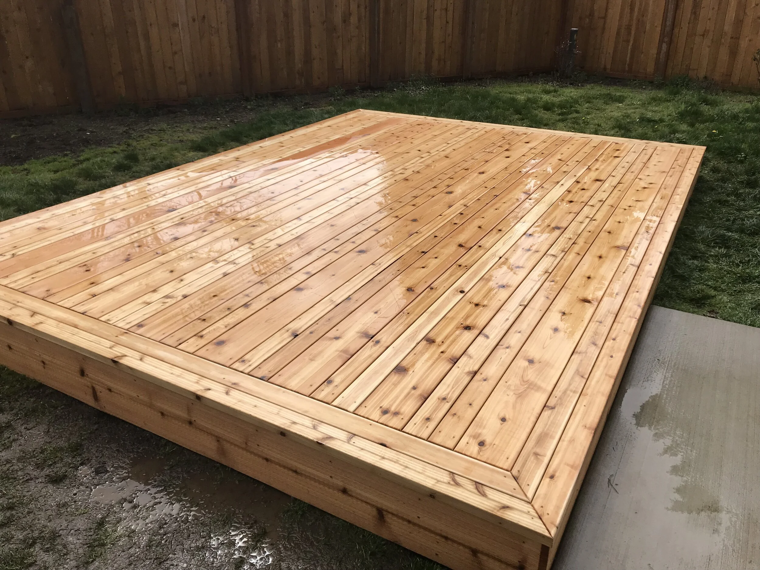 A newly built wooden deck with a rectangular shape, situated in a backyard with a grass lawn and a wooden fence in the background.