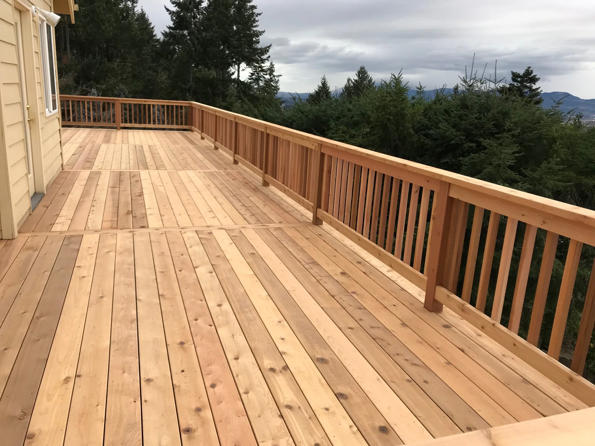 A wooden outdoor deck with a railing, overlooking a forest with evergreen trees and mountain in the distance under a cloudy sky.