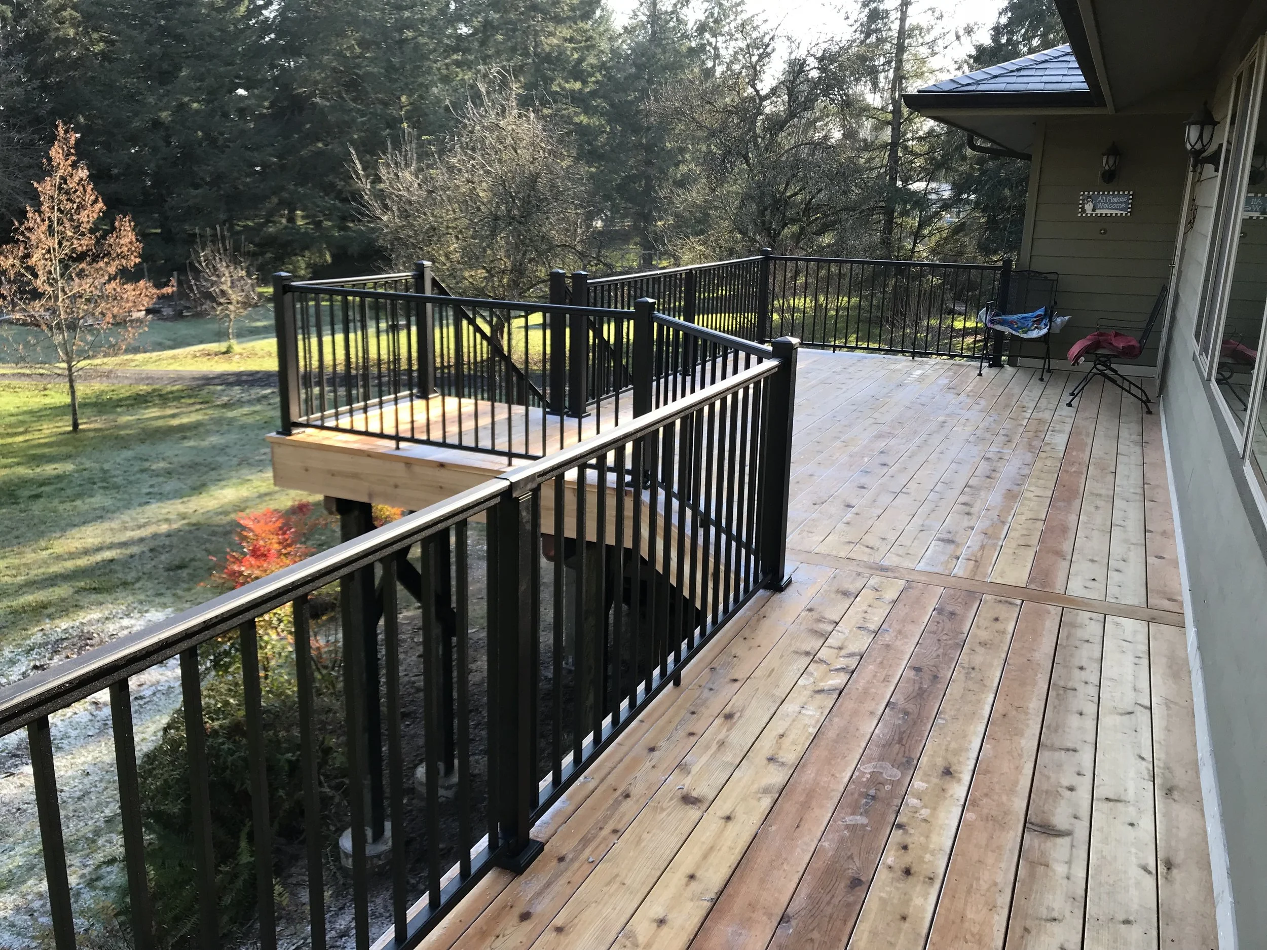 A wooden deck with black metal railings attached to the back of a yellow house. There is a small chair with blankets and a pink jacket on it. The house has siding and a window, with a sign that says 'All Things Welcome'. The yard below has grass, tre